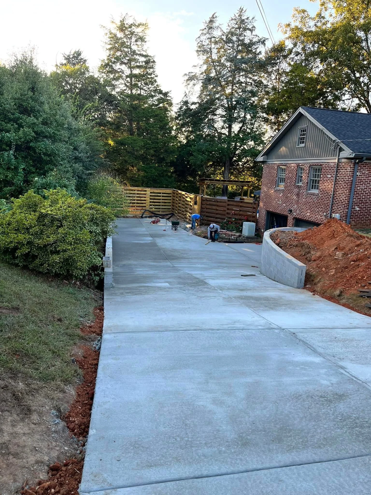 View of a freshly poured concrete driveway with a curved edge on the right, next to a brick house with gray siding and windows, a wooden fence under construction in the background, and a worker kneeling on the driveway.
