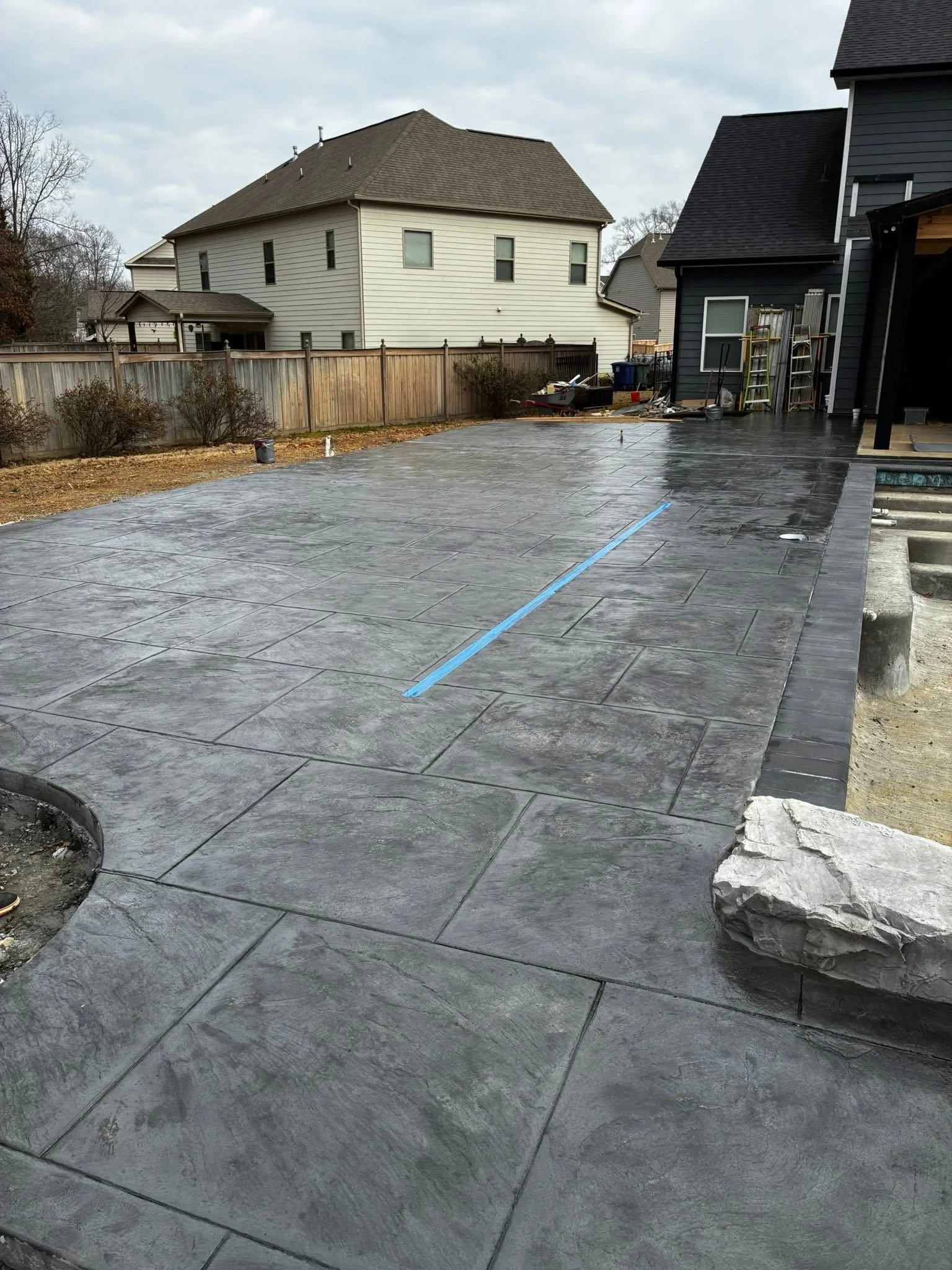 View of a backyard patio with newly finished dark gray stamped concrete, neighboring houses, and a wooden fence.