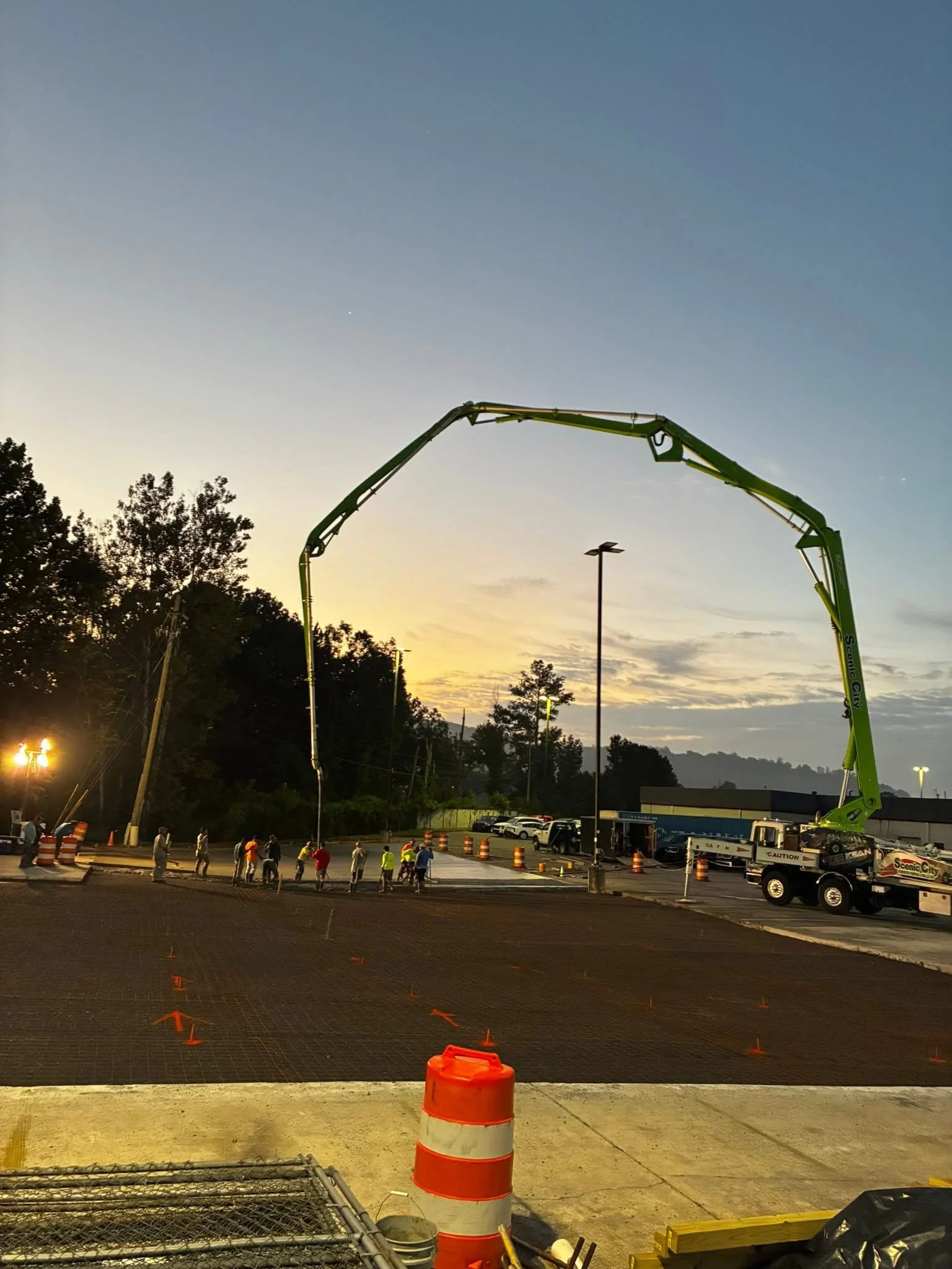 A construction site during early evening with a large green crane arm extending over the area. Workers are standing on the ground, and orange traffic cones mark the construction zone. In the background, there are parked cars, trees, and a building.