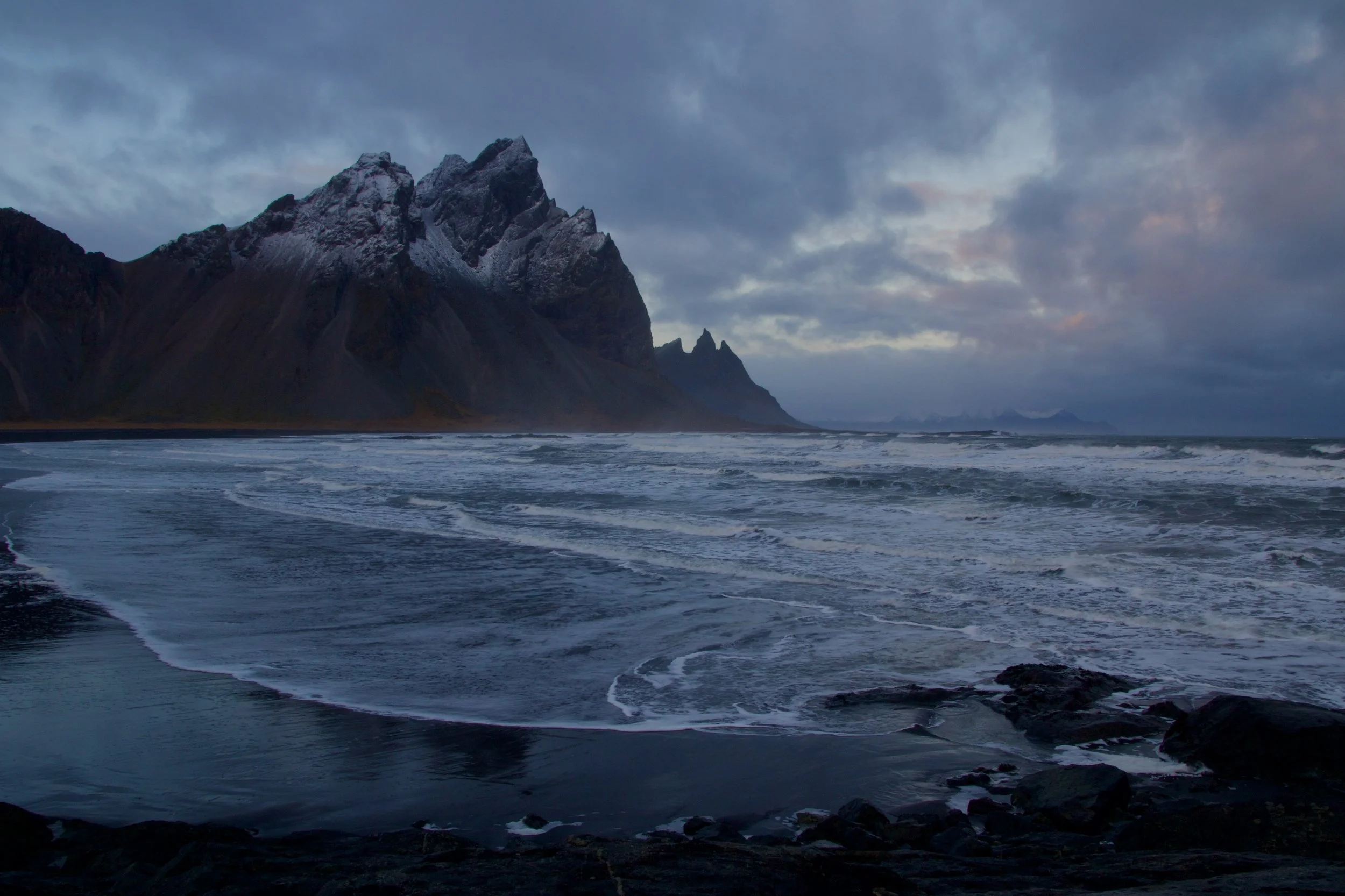 Snow-capped mountains beside a cloudy sky with a rocky shoreline and ocean waves in the foreground.