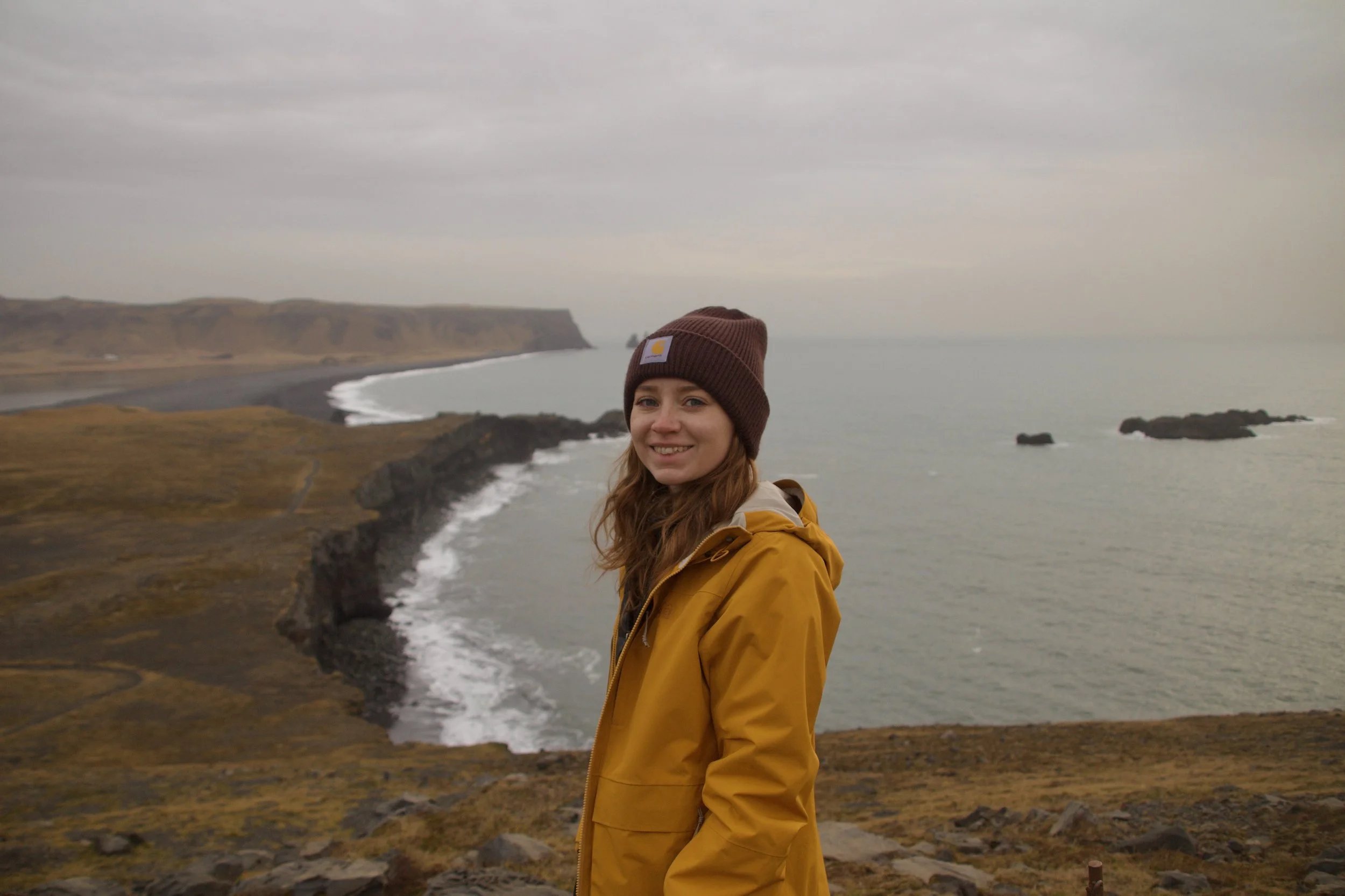 A woman wearing a brown beanie and yellow jacket standing near a coastal cliff with ocean waves and a distant shoreline under an overcast sky.