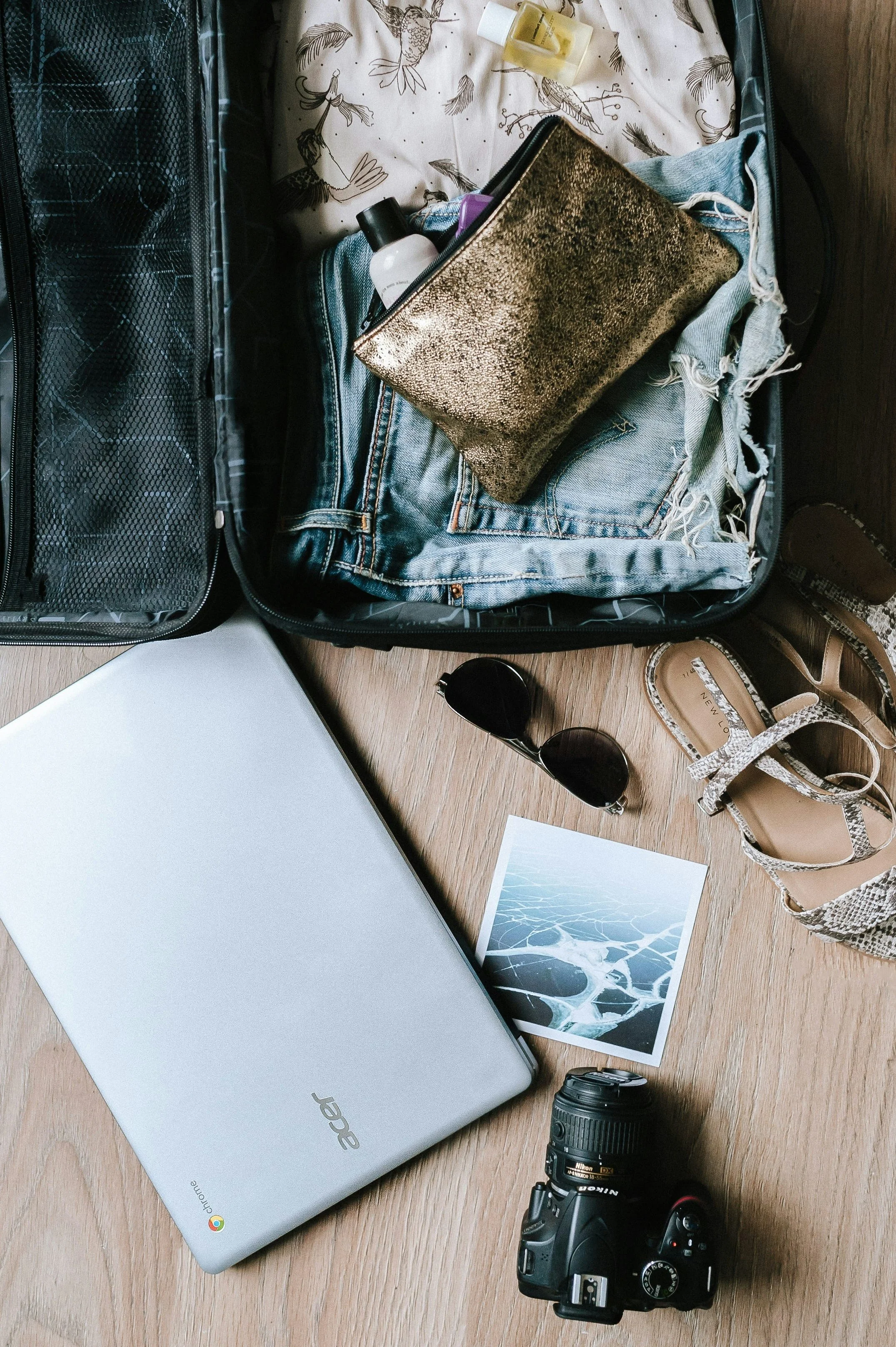 Open suitcase containing clothes, toiletries, and a gold clutch bag, placed on a wooden floor next to a silver Chromebook, a camera, a pair of sunglasses, and a printout of a photo of ice or frozen water.