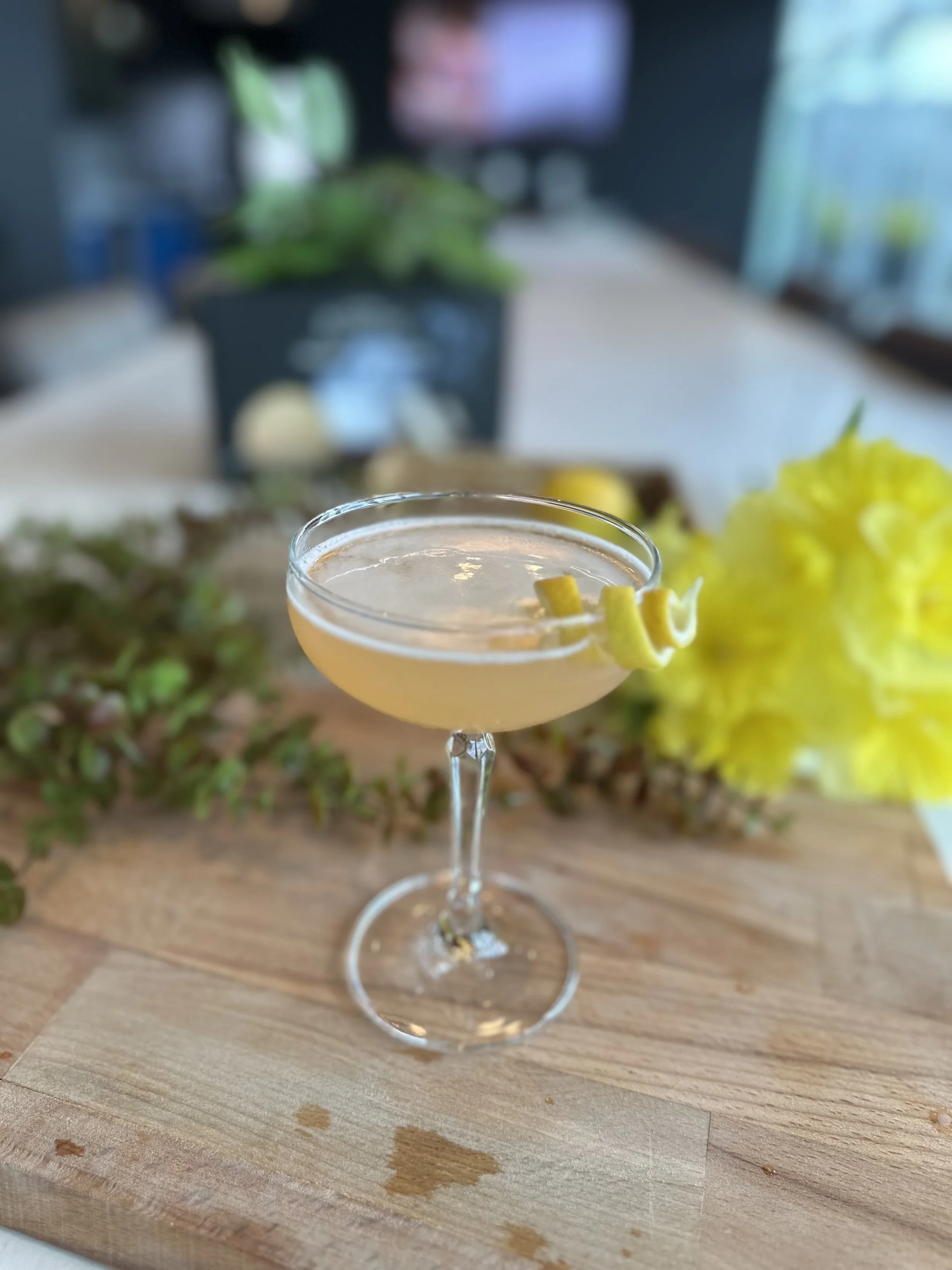 A cocktail in a coupe glass garnished with lemon peel, placed on a wooden surface with a blurred background of greenery and indoors.