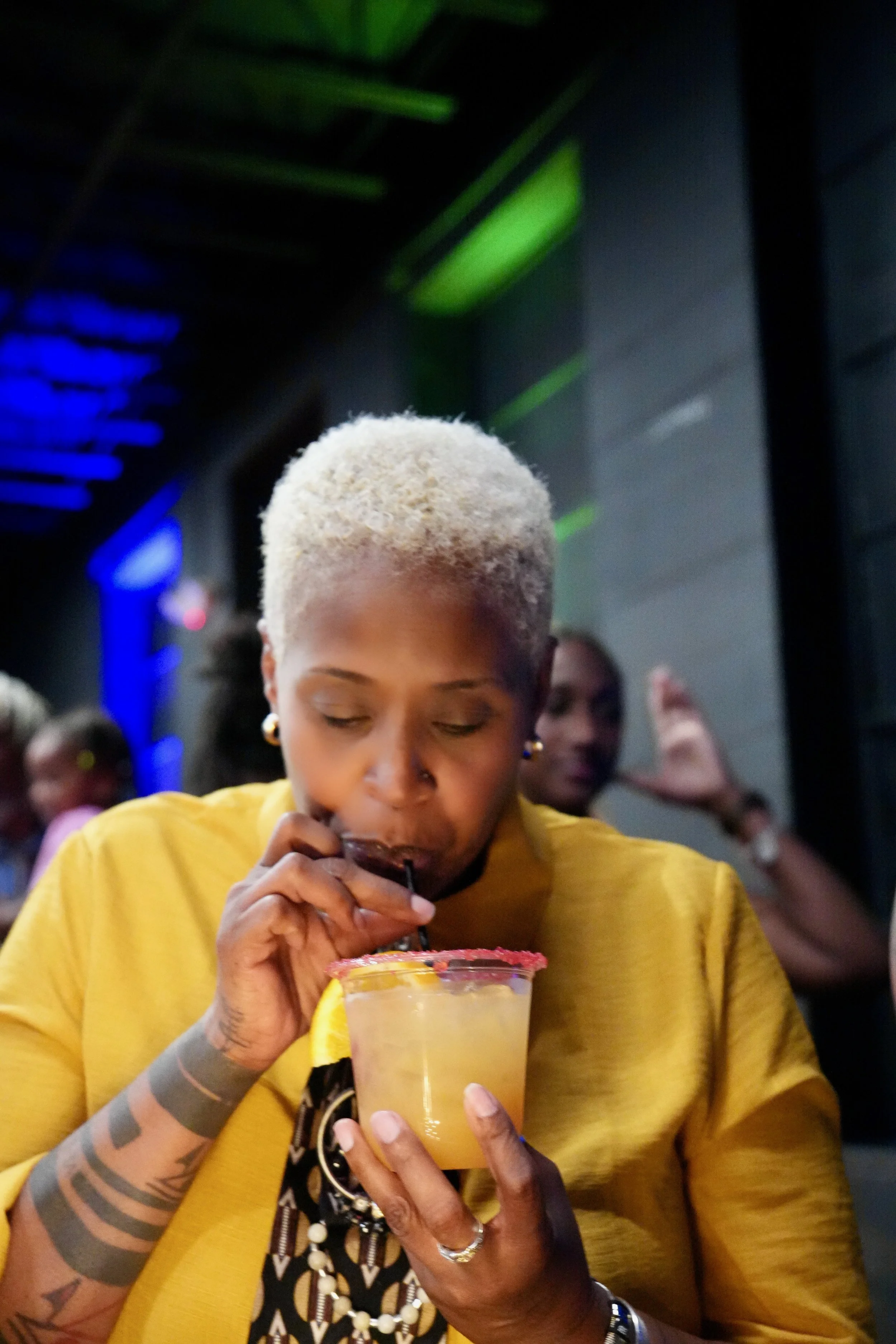 Woman with gray-blonde hair, wearing a yellow shirt, drinking a beverage with a lemon wedge garnish, in a dimly lit setting with other people in the background.