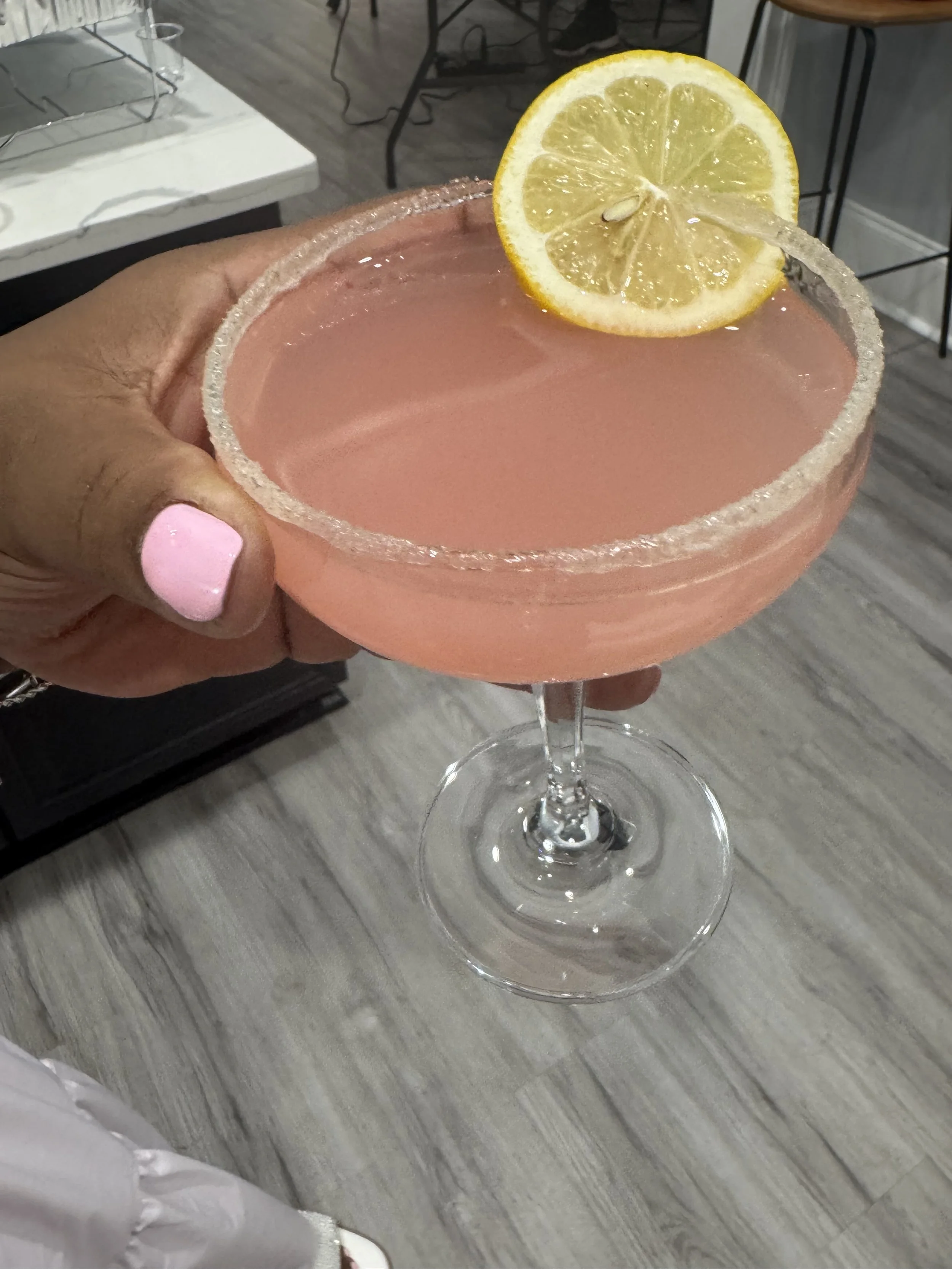 A hand holding a pink cocktail glass with a salted rim, garnished with a slice of lemon and a straw, sitting on a table.