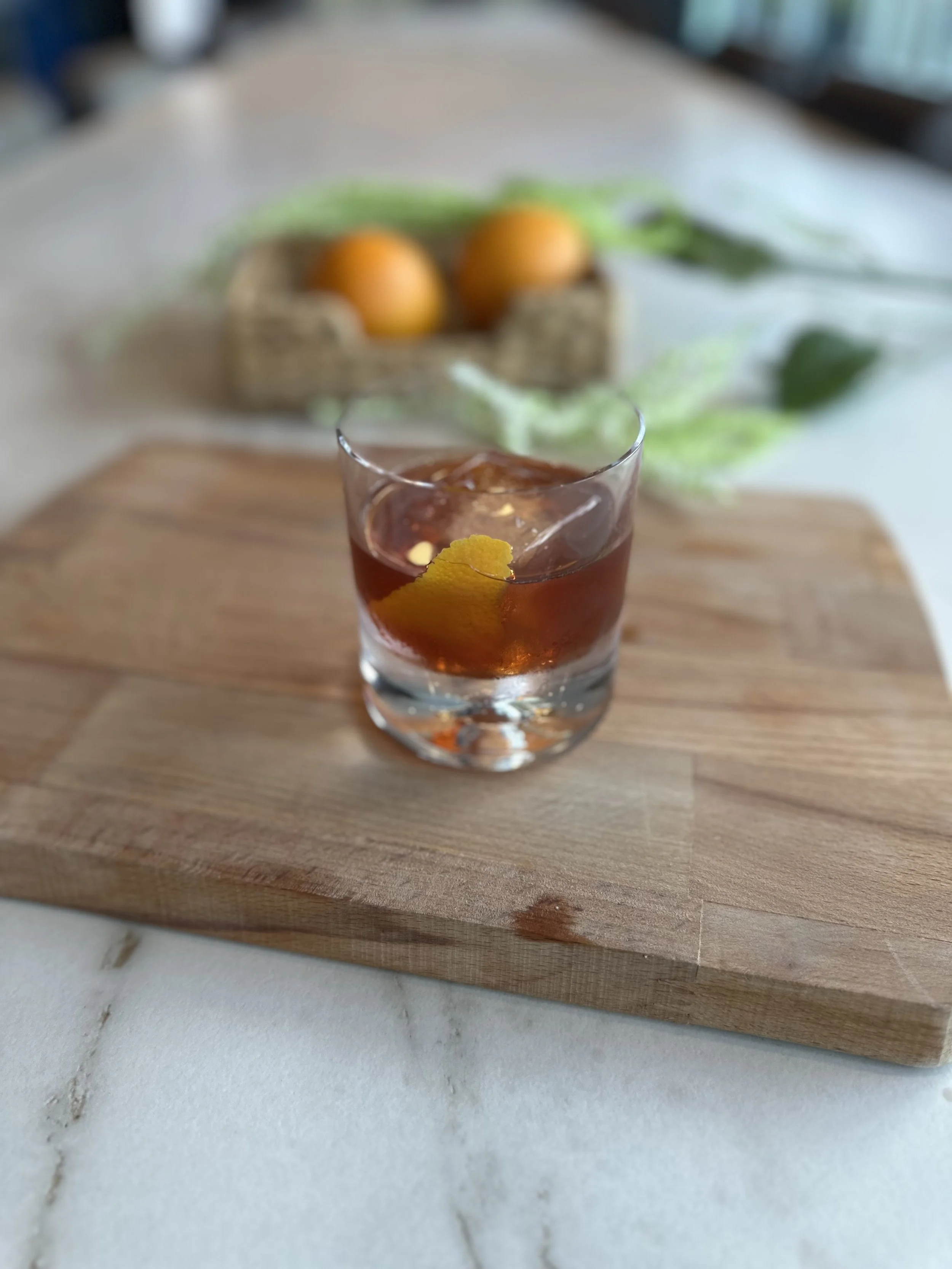 A glass of tea with lemon on a wooden board, with a blurred background of oranges and green leaves.