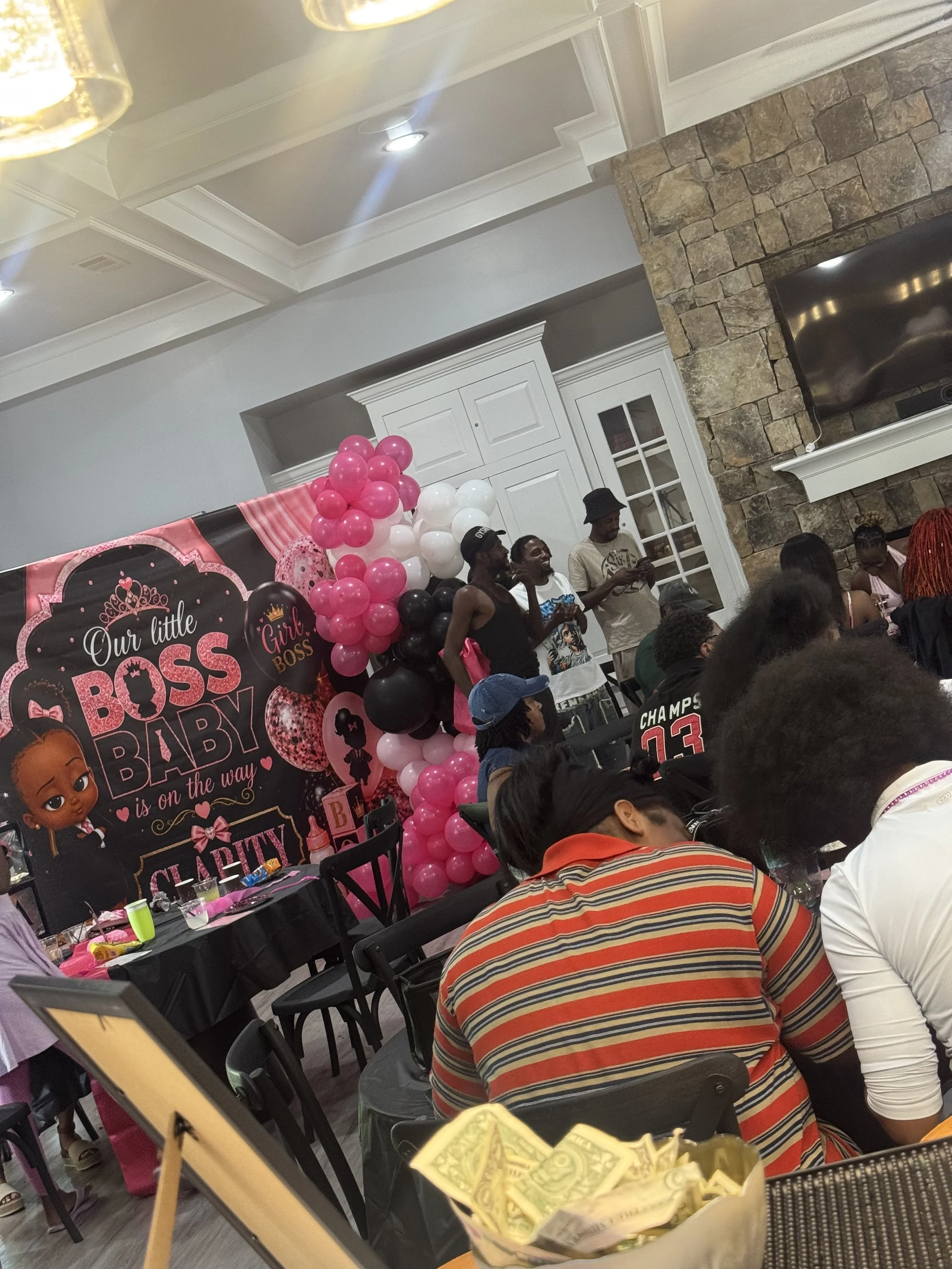 Indoor celebration with people gathered around tables, pink, white, and black balloons, and a backdrop with the words 'Our Little Girl Boss Baby is on the way'.