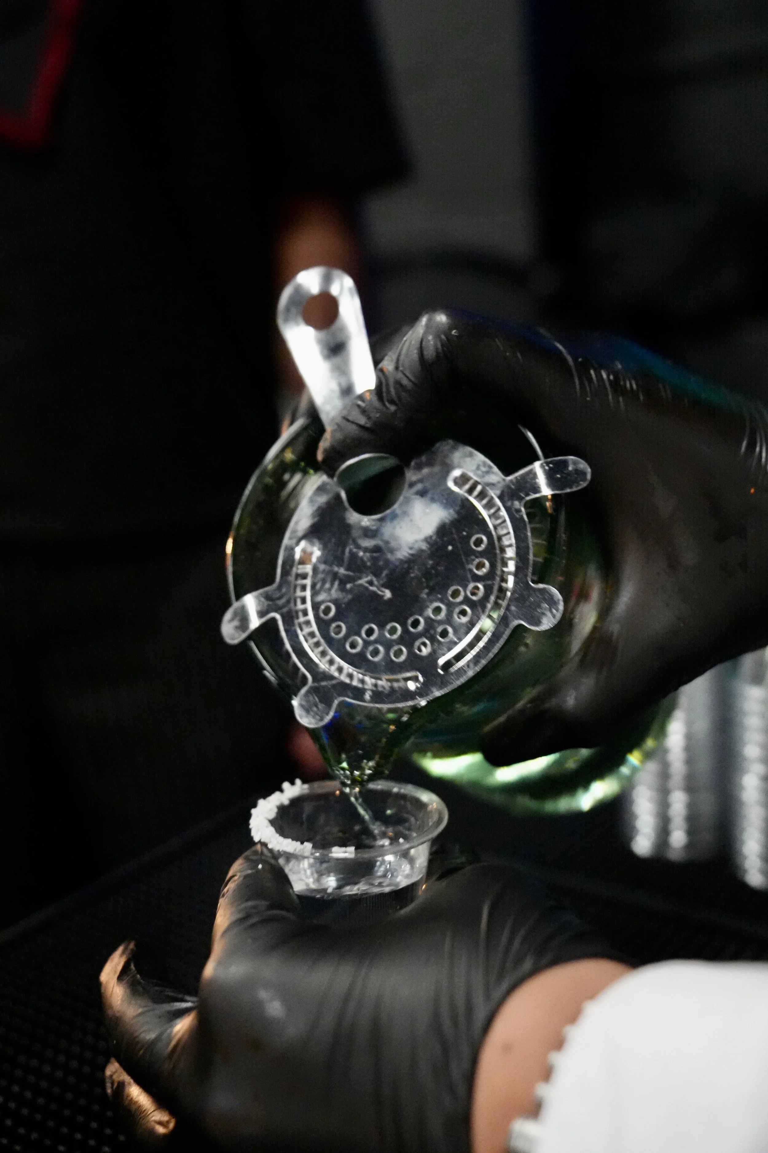 A hand wearing a black glove is pouring a drink from a bottle into a glass.