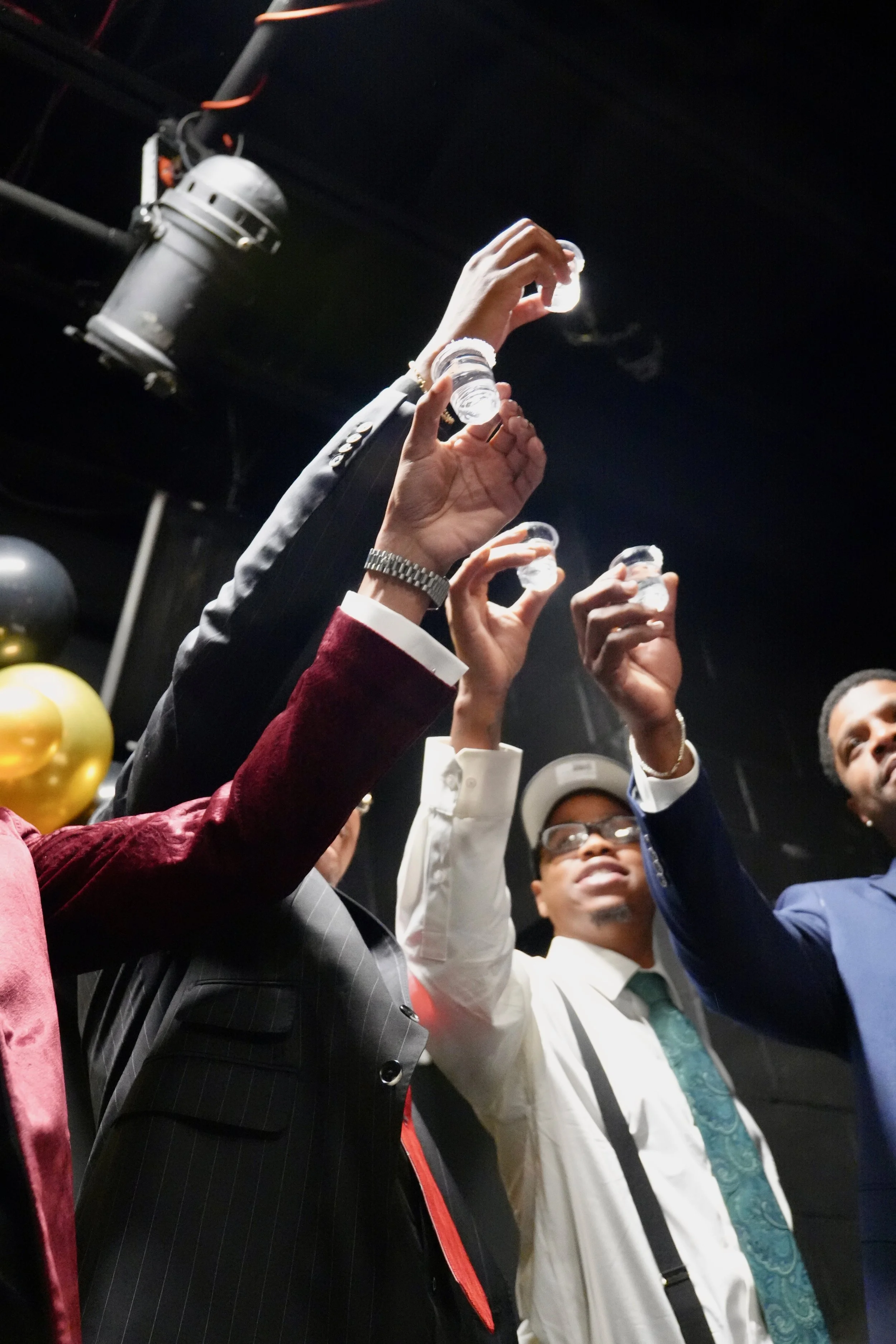 Group of people raising their hands, some holding small cups, in celebration or greeting, indoors with black ceiling and balloons visible in the background.