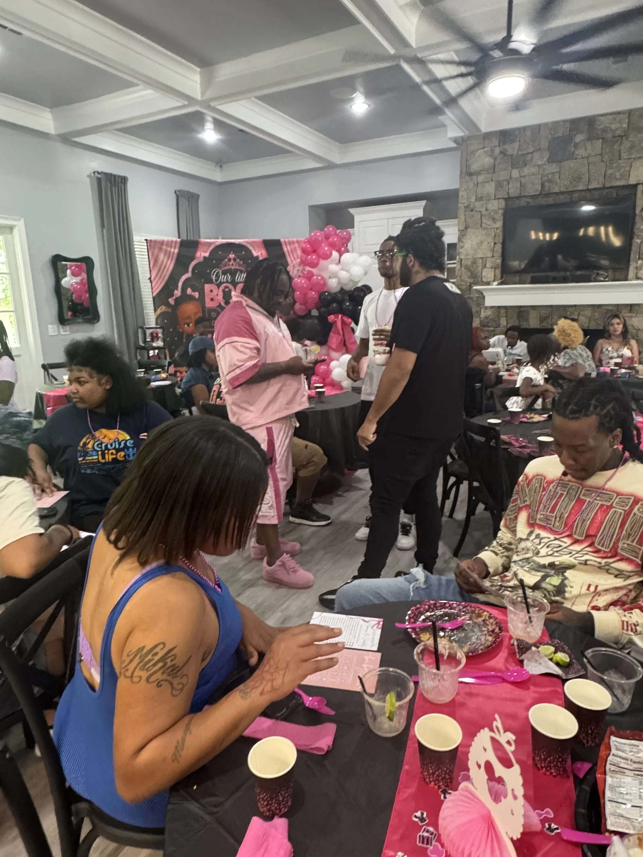 A woman with a tattoo on her arm sitting at a table with pink paper decorations, drinks, and party supplies, reading a card. The table has birthday-themed decorations and cups filled with drinks. In the background, there are other guests, a balloon decoration, and a banner that reads 'Our Little Boss Baby'.