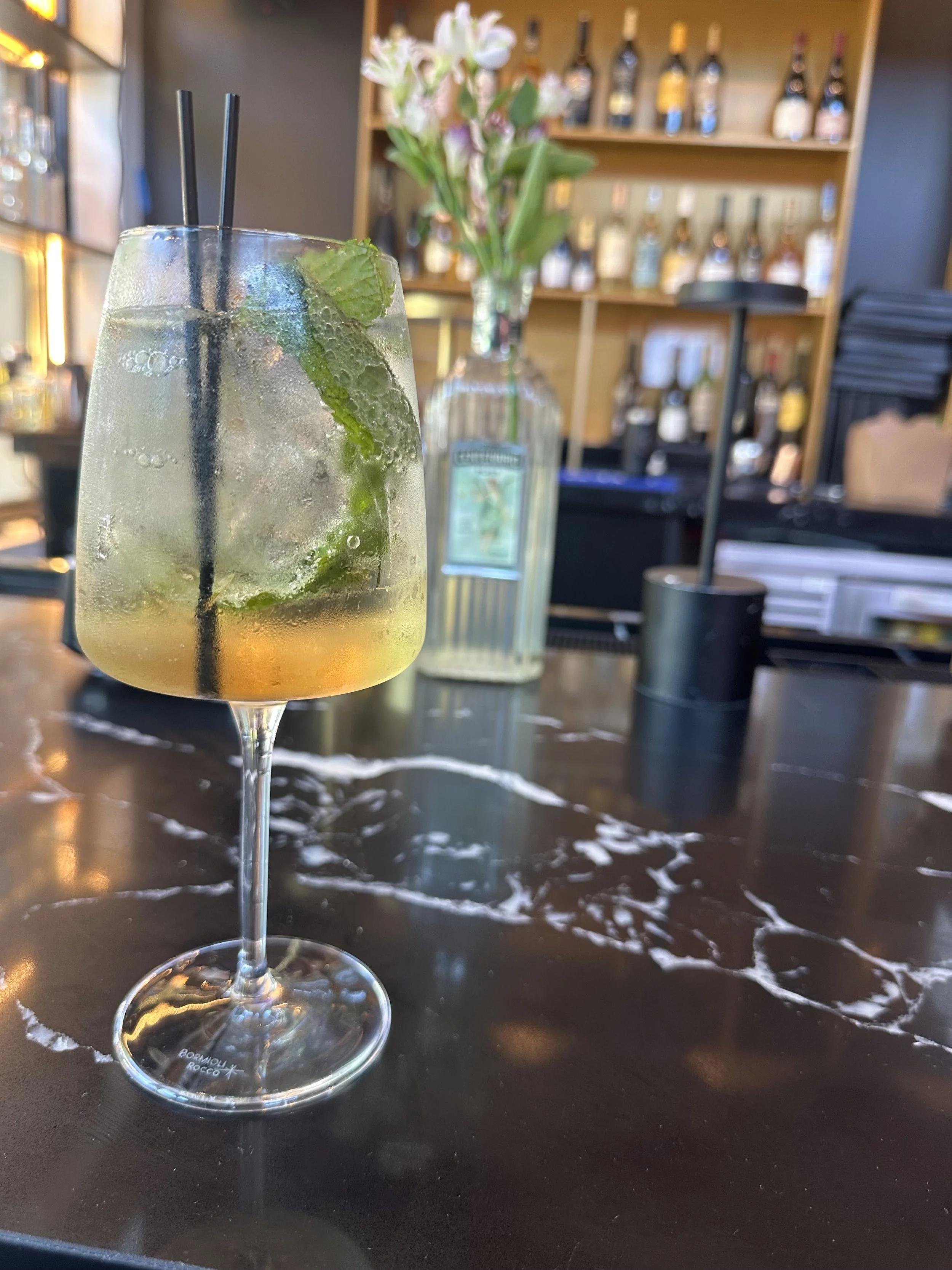 A cocktail glass filled with a light-colored beverage, ice, fresh mint leaves, and two black straws, on a dark table with white marbling.