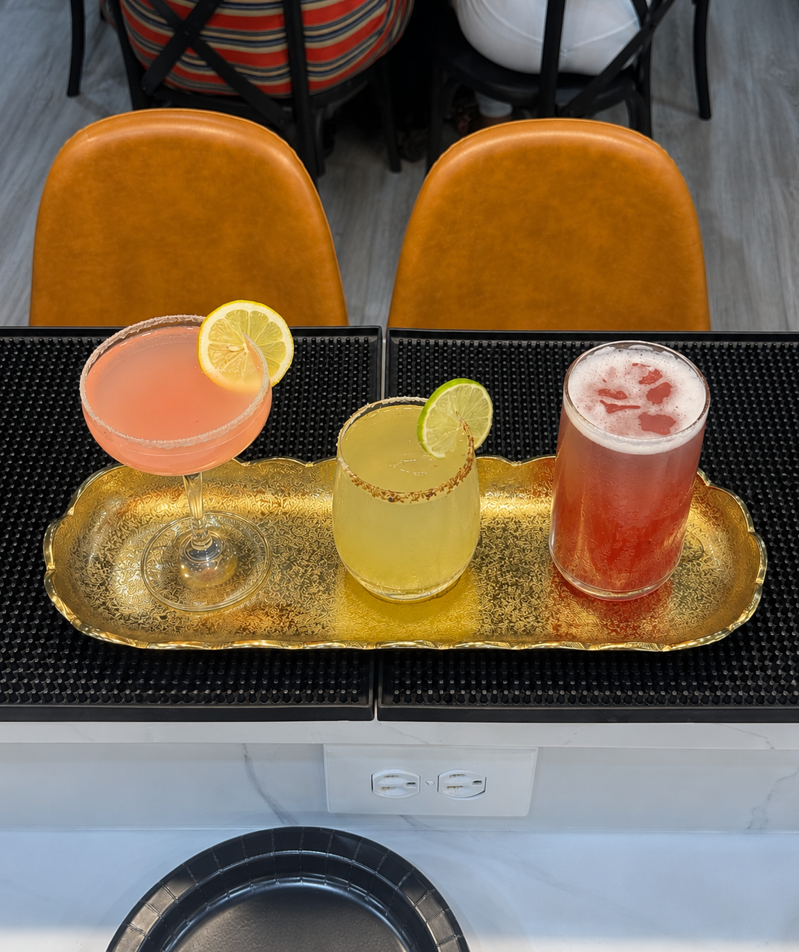 Three cocktails placed on a gold decorative tray in front of two orange chairs at a dining table.
