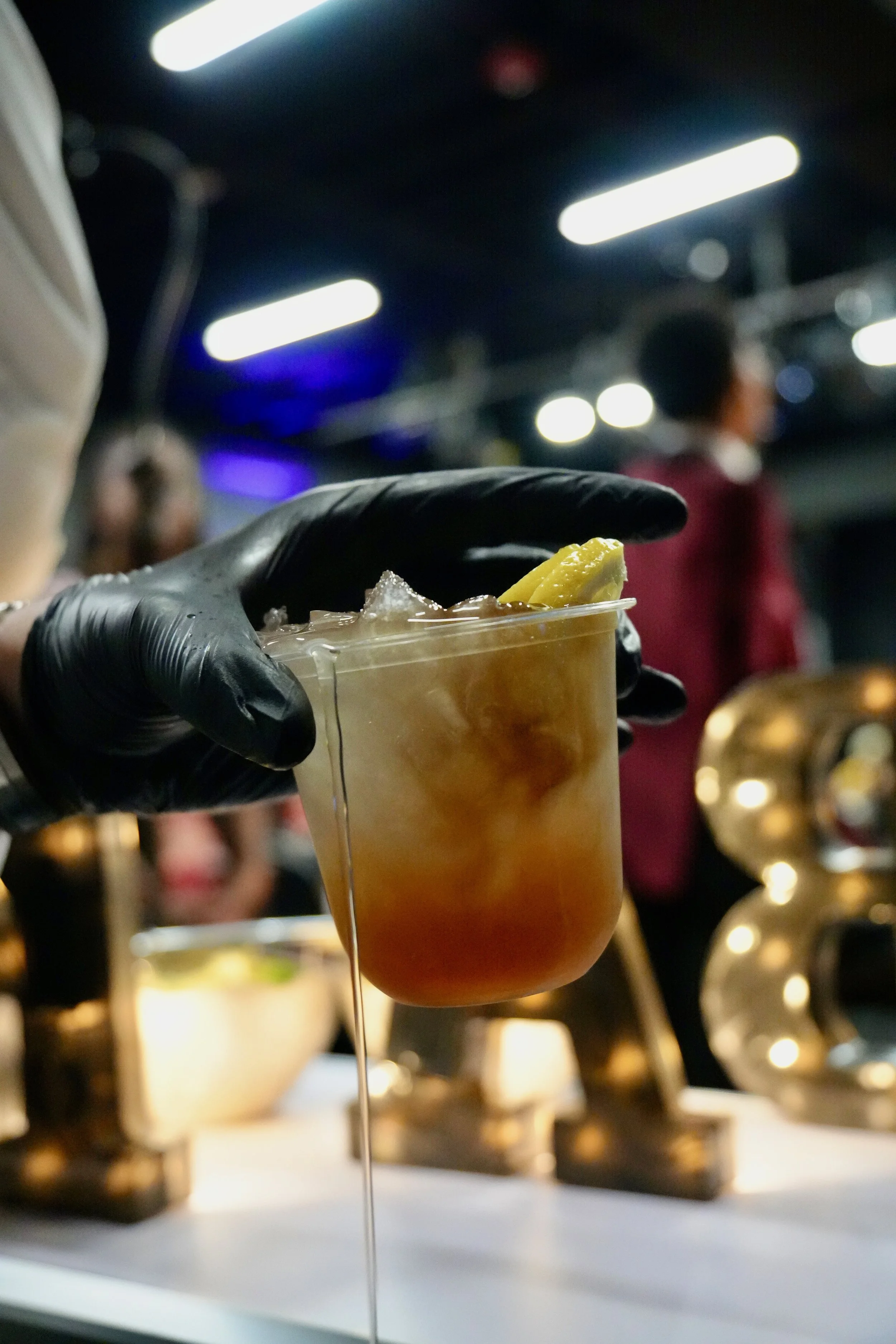 A person wearing a black glove holding a transparent cup filled with a brown beverage and a lemon wedge garnish, with other people and blurred lighting in the background.