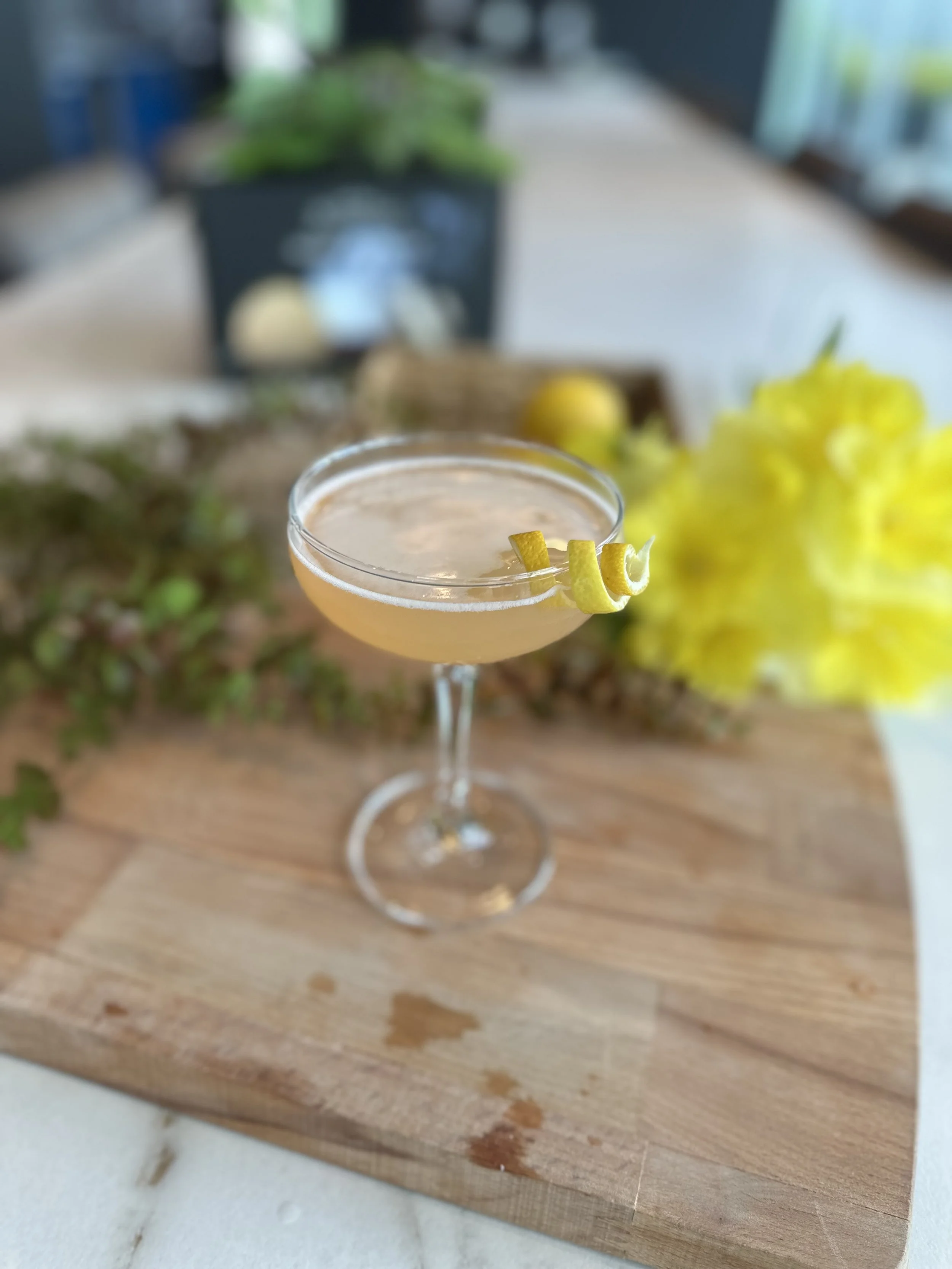 A cocktail glass with a yellow lemon twist garnish on the rim, placed on a wooden surface with greenery and blurry interior background.