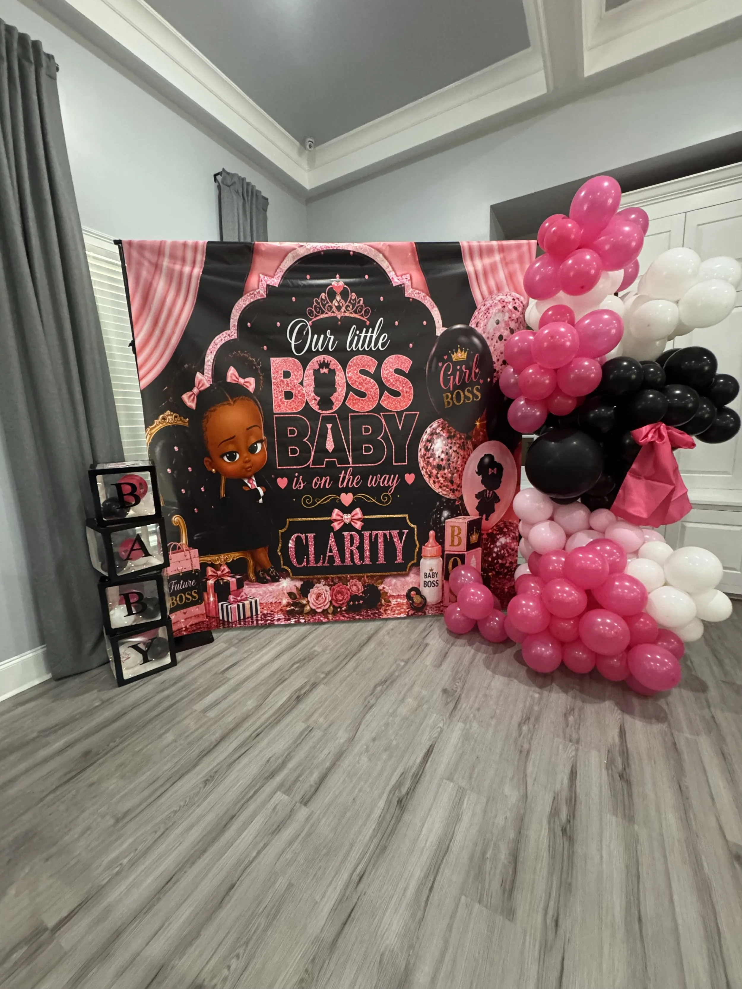 Decorative birthday display featuring a pink, black, and white balloon arrangement next to a backdrop with a little girl princess theme, with pink and black balloons, gift boxes, and a sign that says 'Our little boss baby is on the way'.