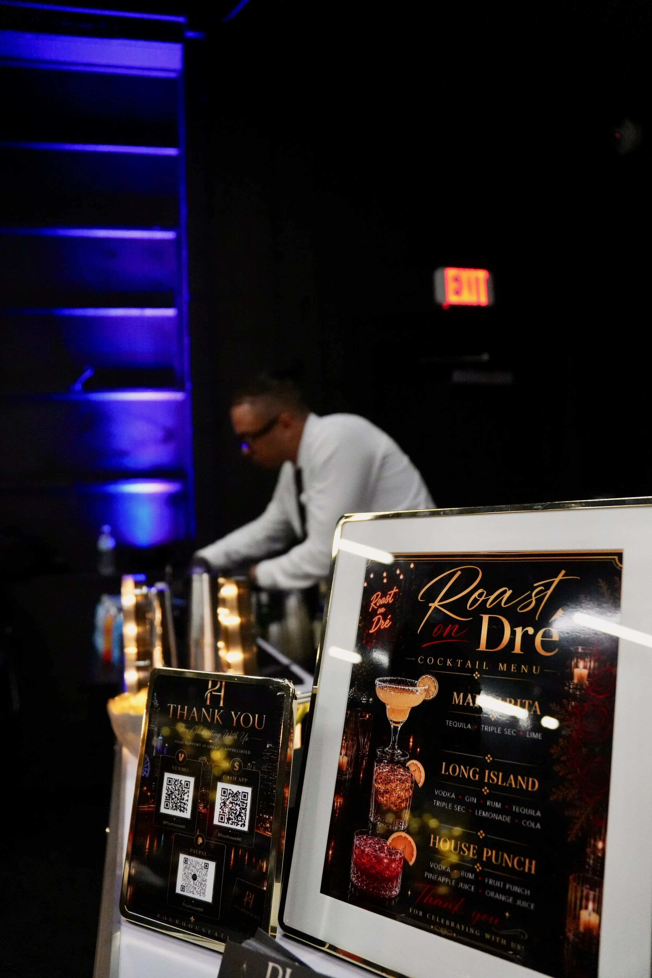 A dimly lit bar or nightclub with a man in a white shirt behind the counter. Two cocktail menus are prominently displayed, featuring drinks like Long Island and House Punch, with a blue-lit shelf on the left and an illuminated Exit sign on the wall.