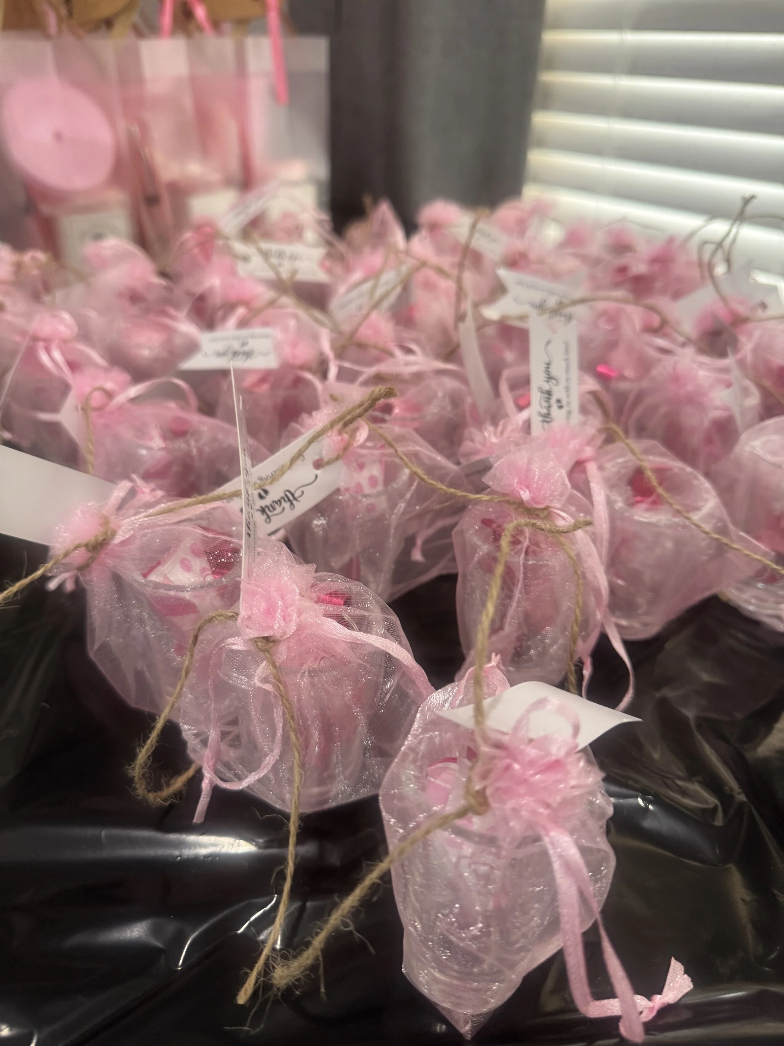 Pink organza favor bags tied with pink ribbons, arranged on a black surface in front of a window with white blinds.