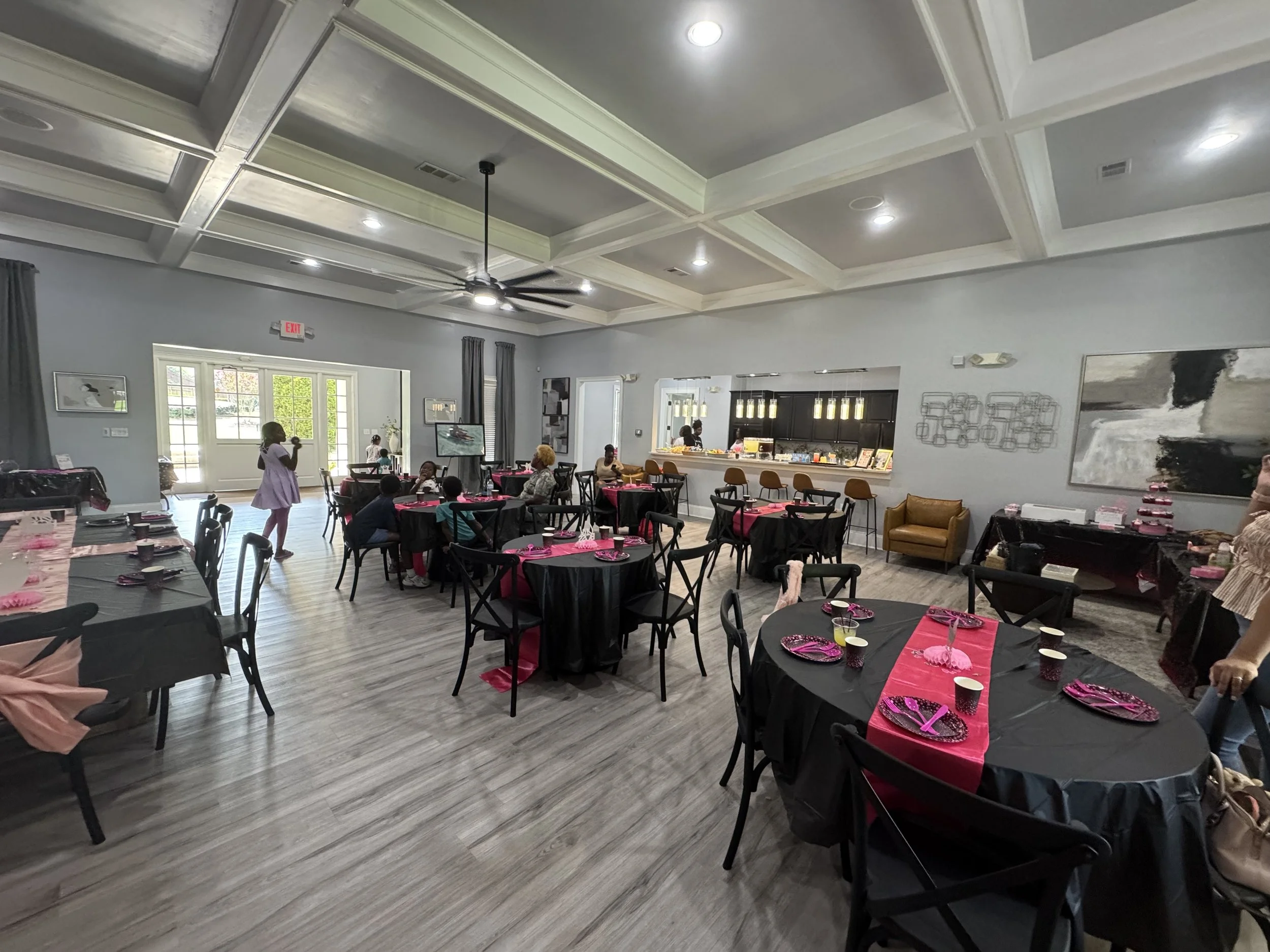 A large banquet room is decorated with pink and black table settings for a celebration. There are children and adults seated at round tables, with some standing and talking. The room has a high ceiling with recessed lighting and a ceiling fan, gray w