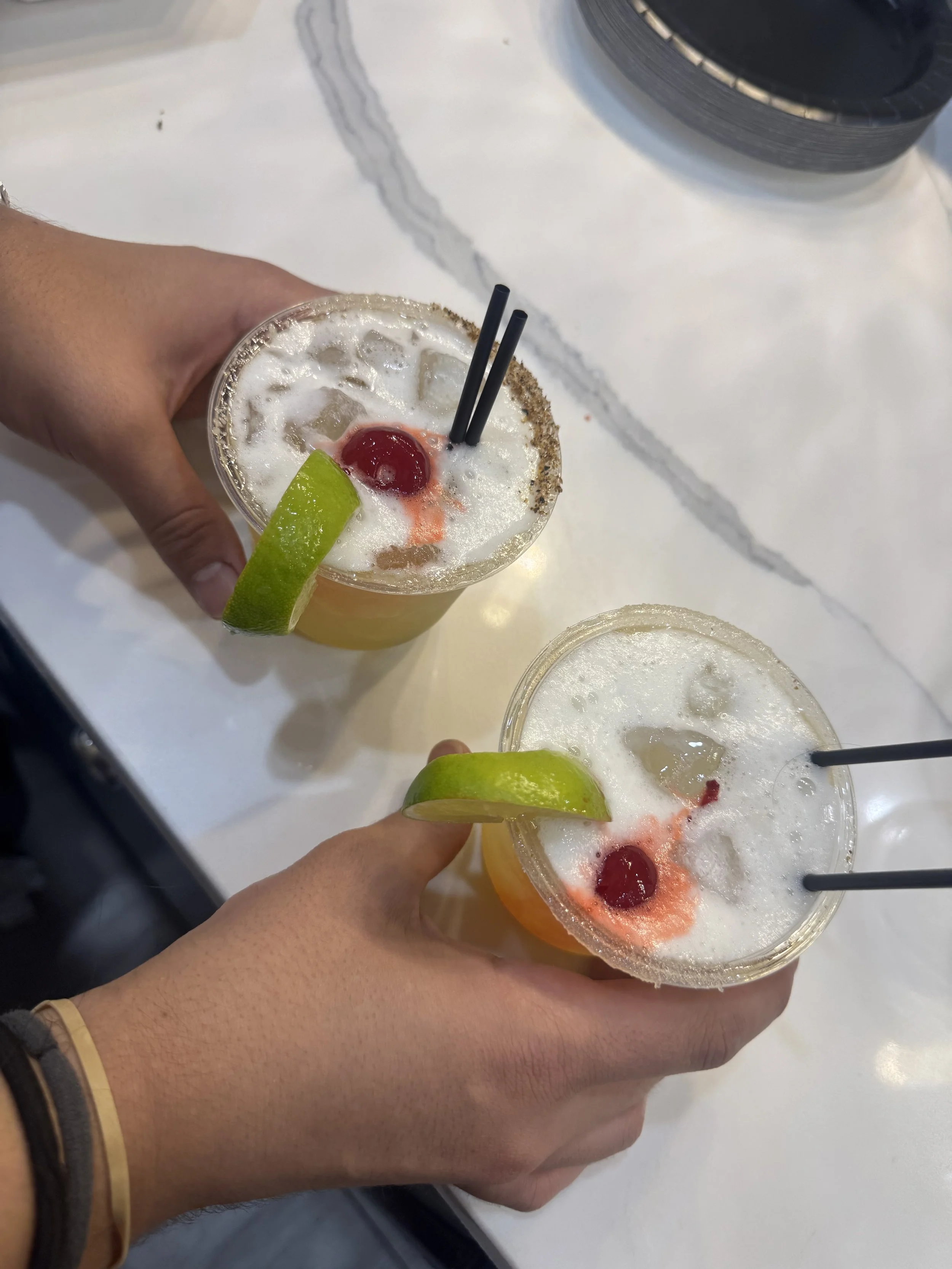 Two hands holding Margarita cocktails with pink rims, lime wedges, and cherries, on a white table.