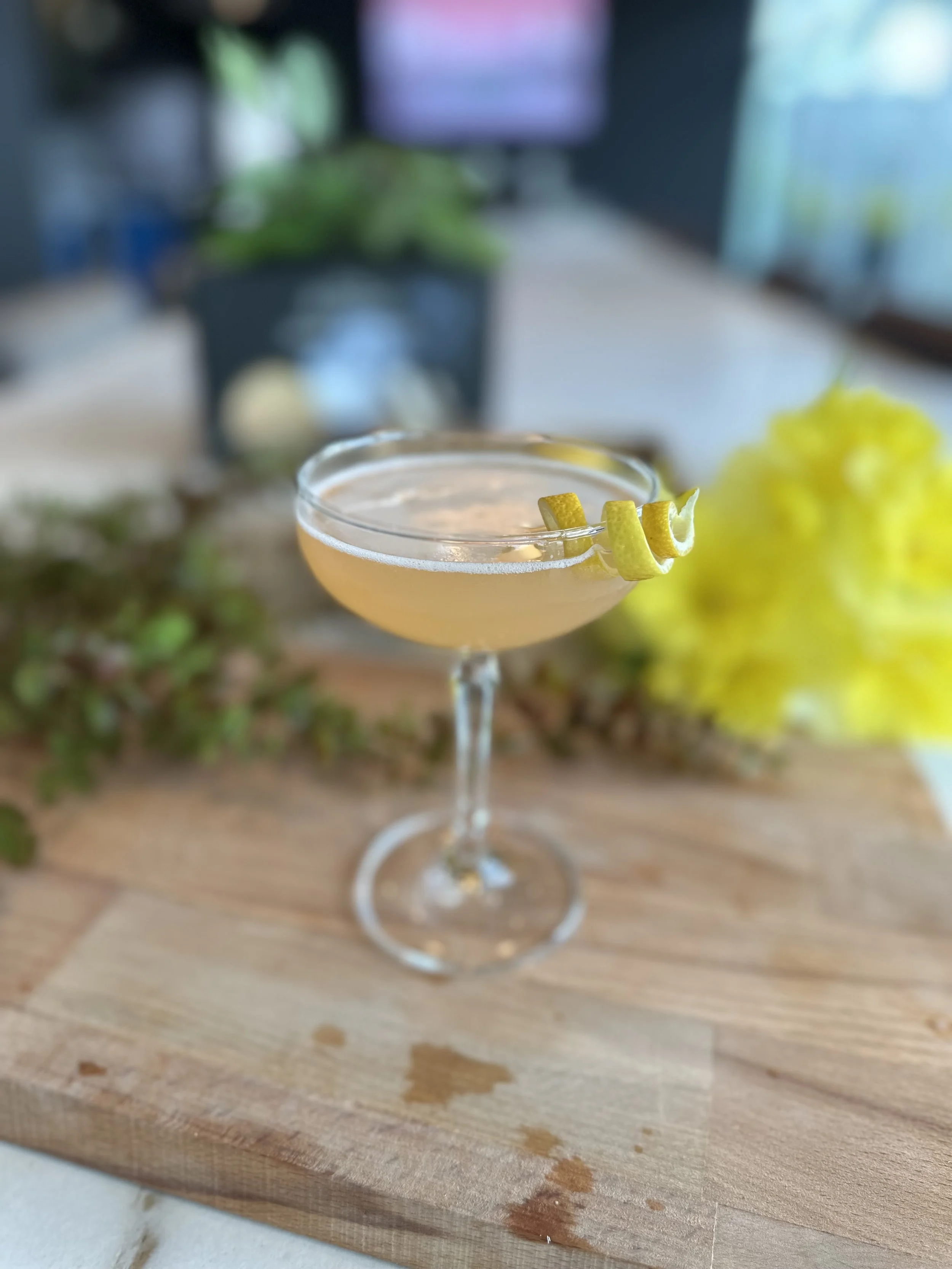 A cocktail glass with a light-colored drink, garnished with a twisted lemon peel, placed on a wooden surface with some wet spots around it. In the background, there is a blurred dark-colored container with a plant, some greenery, and what appears to be a TV or computer screen.