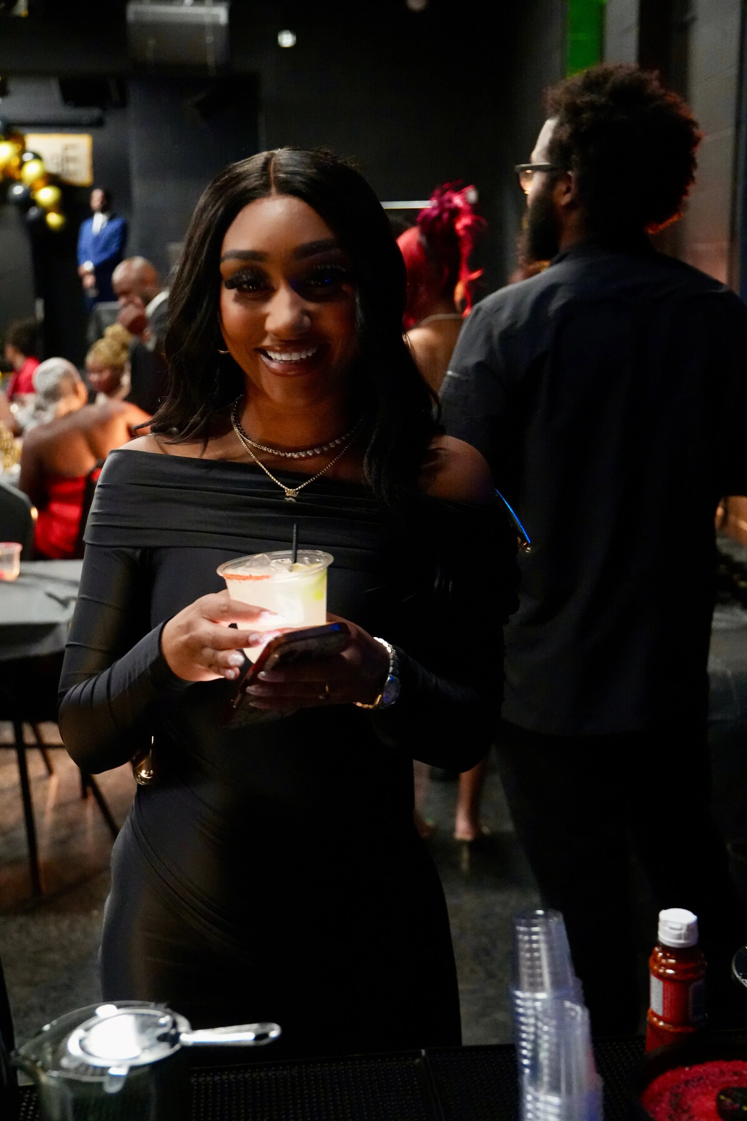 A smiling woman with long black hair holding a drink and a phone at a social event. People in the background are engaged in conversation, and there are balloons and decorations on the wall.