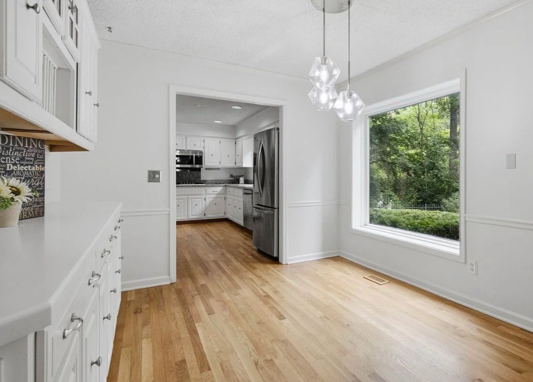Bright white kitchen with large window showing green trees outside, wooden floors, white cabinetry, and a modern light fixture.