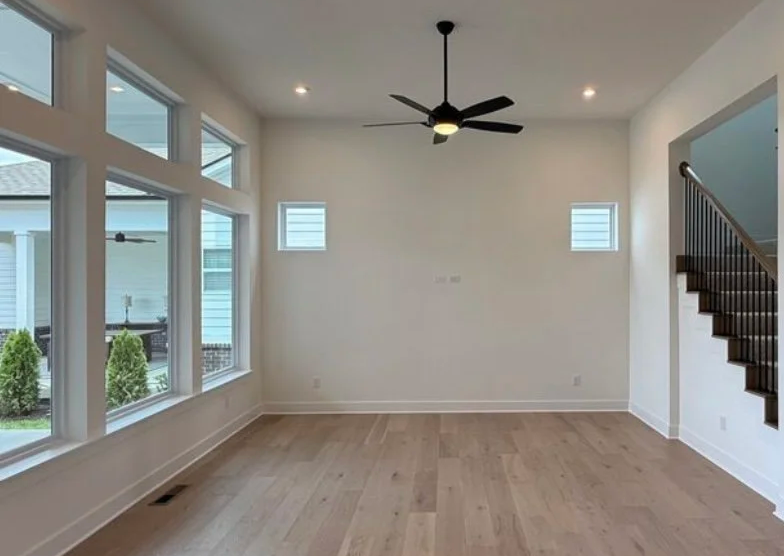 Empty living room with large windows, a ceiling fan, and staircase with dark wood railing on the right.