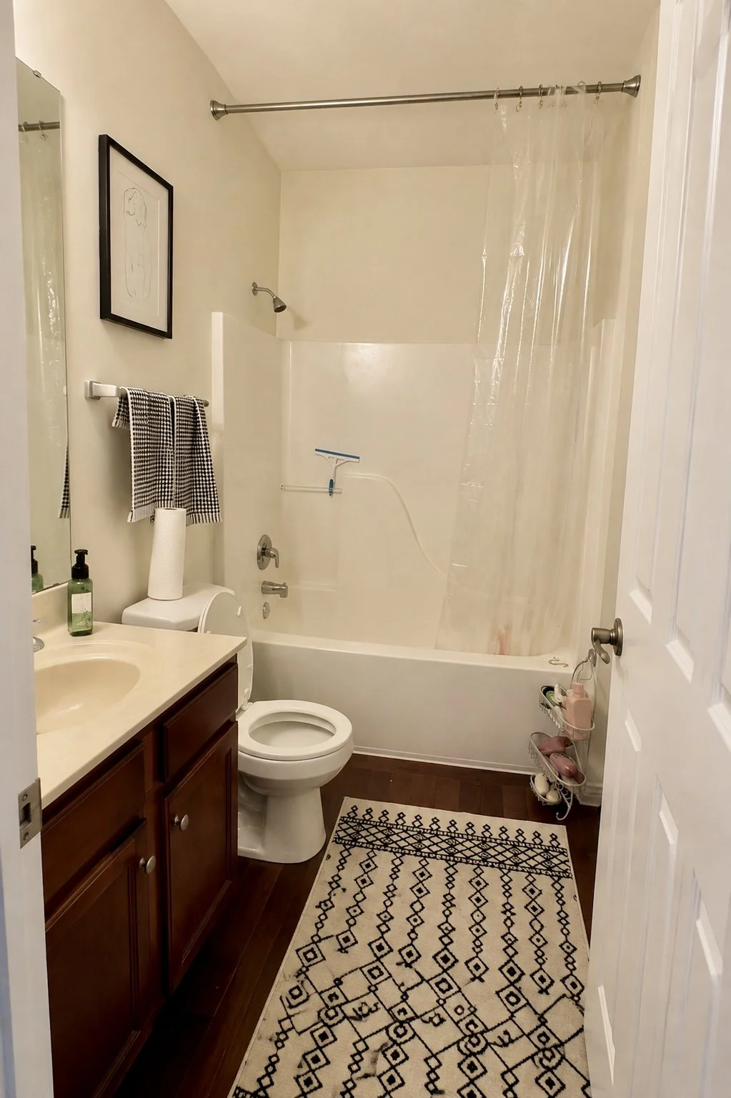 A small bathroom with a bathtub, shower, toilet, sink, and wooden vanity. There is a black and white patterned rug on the dark wood floor, a mirror, and a framed line drawing on the wall.