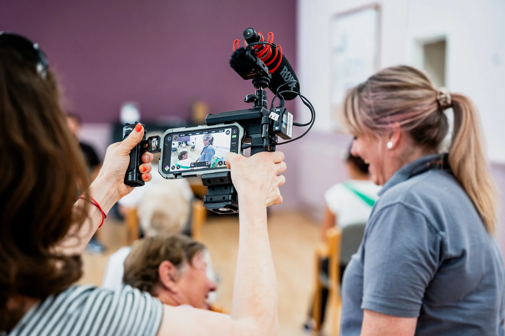 A person operating a camera on a tripod captures a photo or video of a classroom setting as part of media and podcast speaker training.