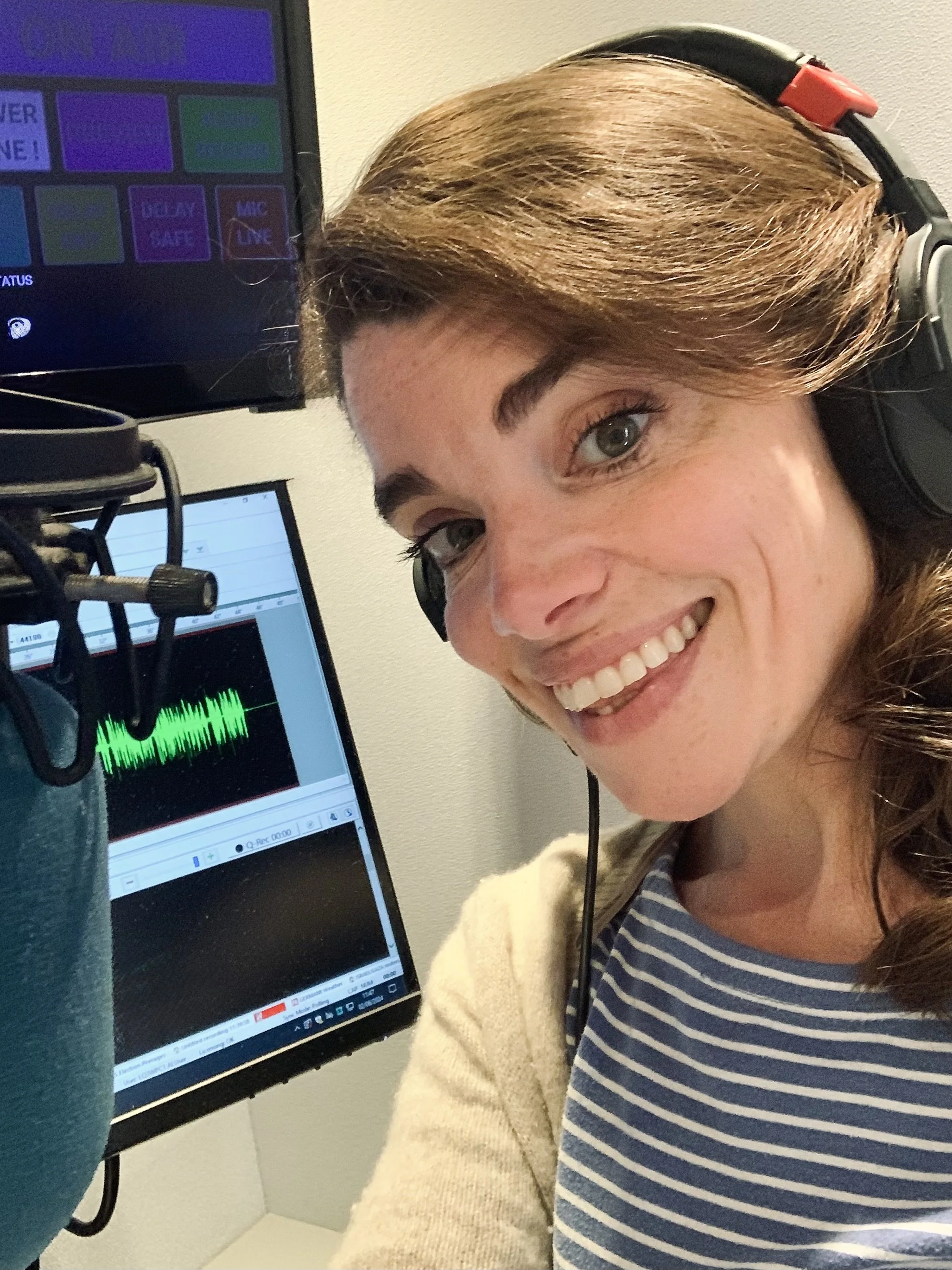A woman with light brown hair, wearing headphones, smiling at a microphone, with a computer screen showing audio editing software in front of her.
