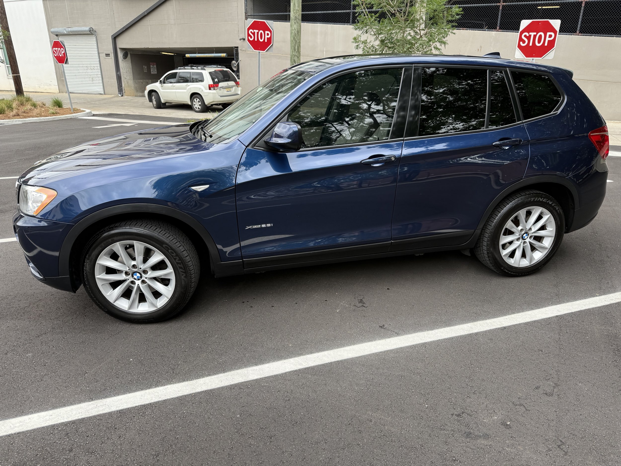 Blue BMW SUV parked in a parking lot with STOP signs, a white SUV near the entrance, and a building in the background.