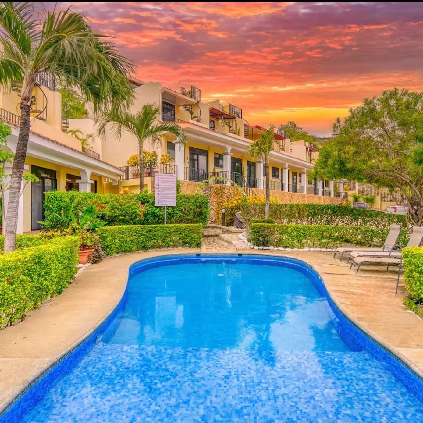 A swimming pool in a resort courtyard with lounge chairs, surrounded by lush greenery, with a sunset sky in the background and multi-story residential buildings.