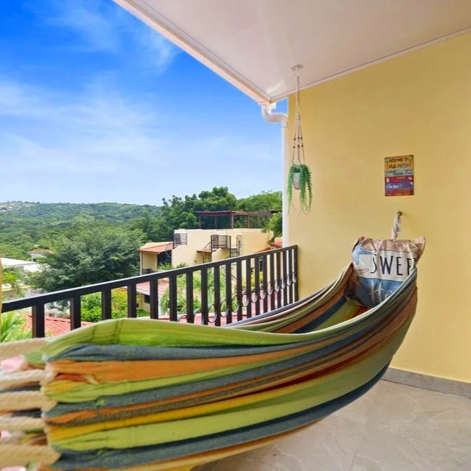 View of a balcony with a colorful hammock, yellow wall, and decorative plants, overlooking a green landscape under a blue sky.
