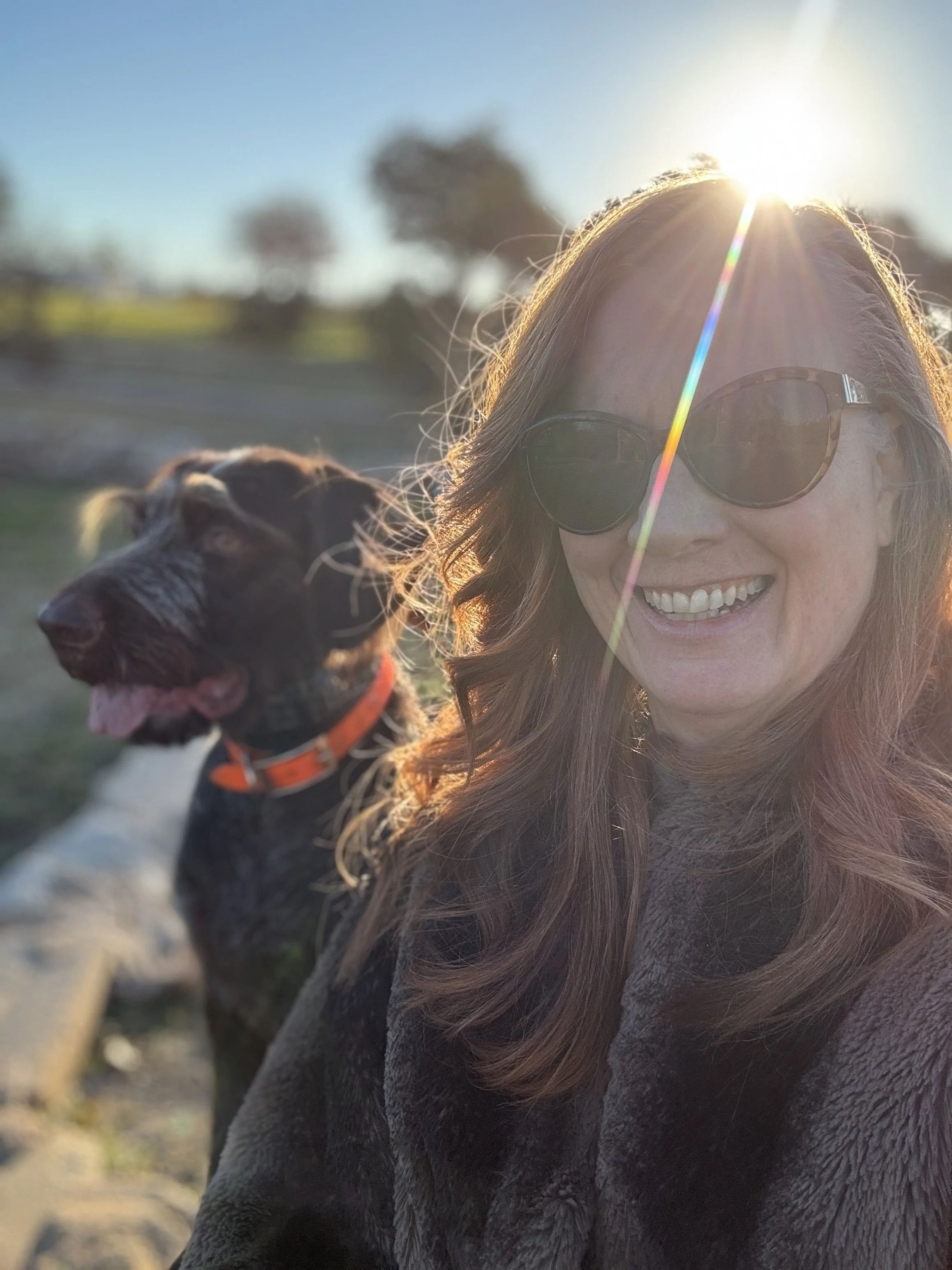 A woman with long red hair wearing sunglasses smiling, outdoors at sunrise, with a brown dog wearing an orange collar nearby. The sun streams down, casting ray across her head.