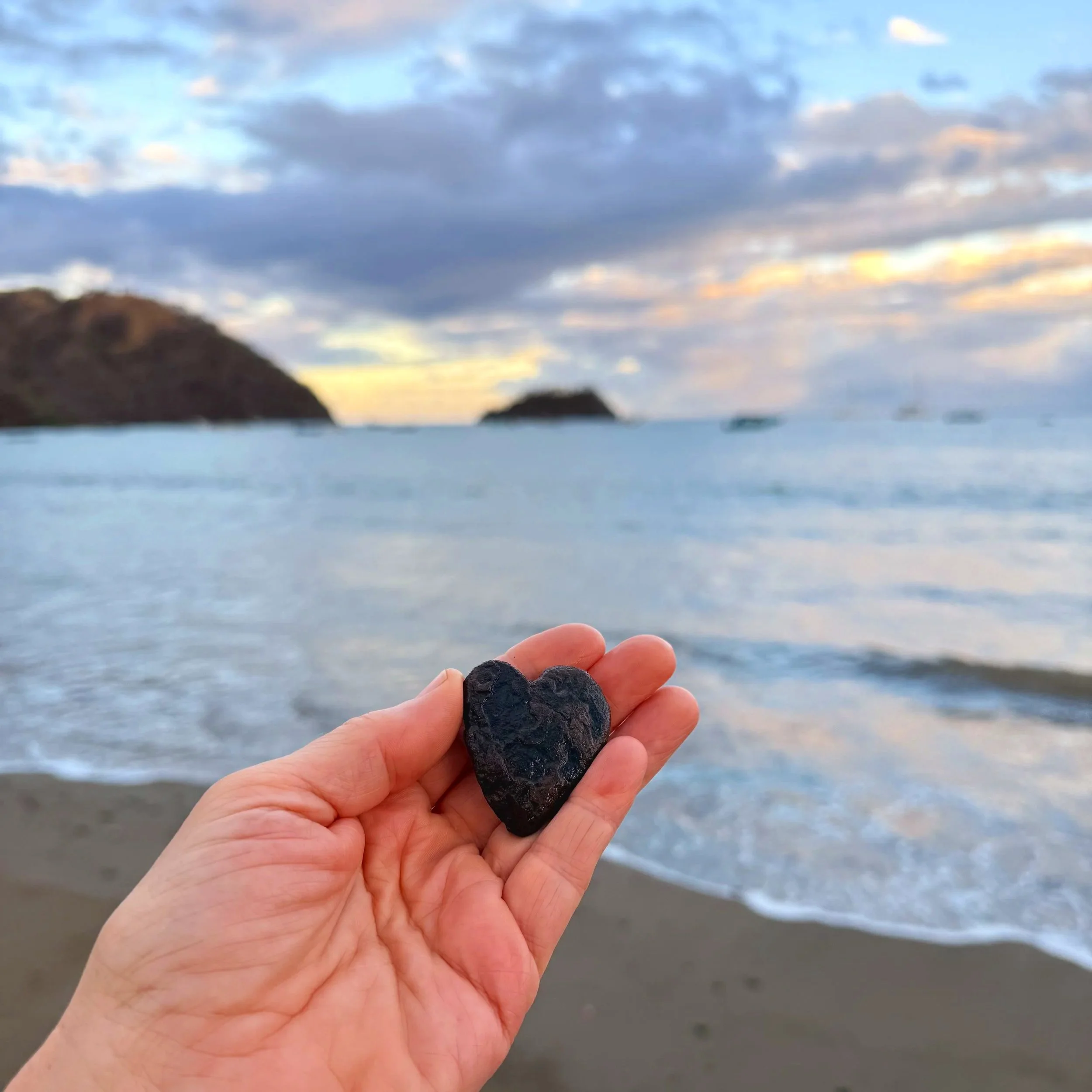 Close-up of a hand holding a small black volcanic rock, with a beach and ocean in the background during sunset.