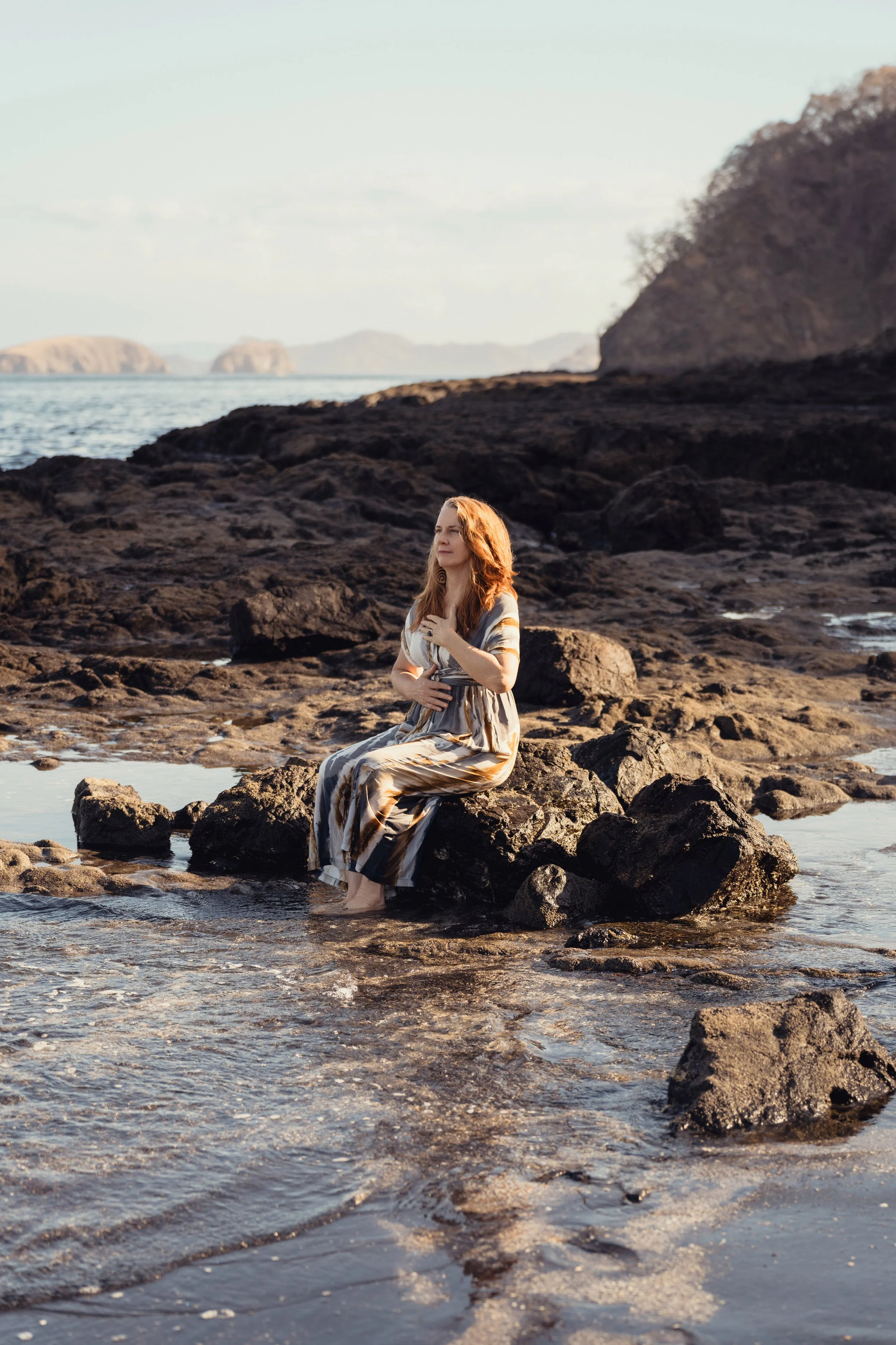 A woman with long red hair wearing a flowing dress sits on rocks at the edge of the ocean with her feet in the water, hand on her chest, gazing into the distance during sunrise.