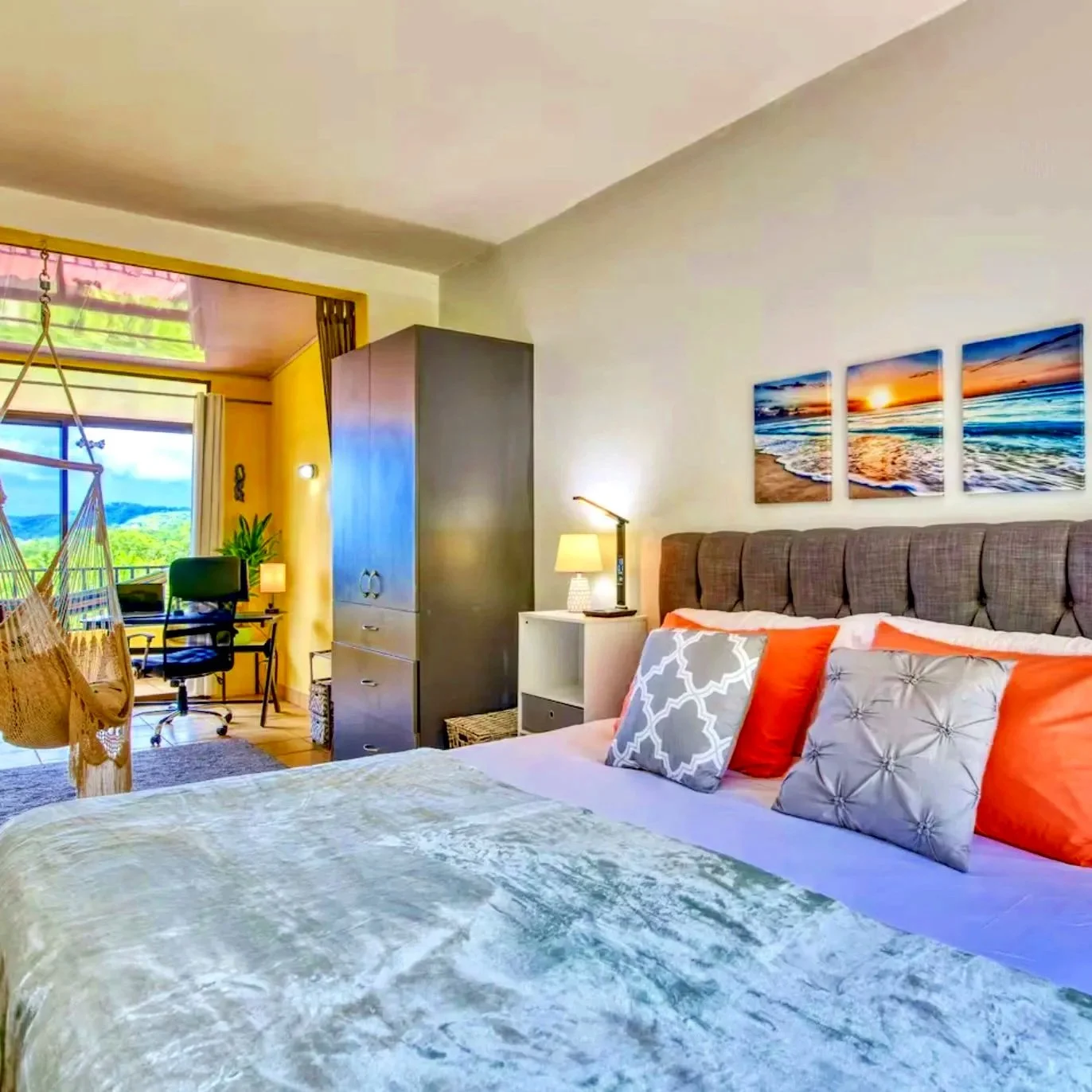 A bedroom with a bed having orange and grey pillows, a white nightstand with a lamp, a colorful triptych artwork of a beach scene, a wardrobe, and a workspace with a desk, chair, and large window showing greenery outside.