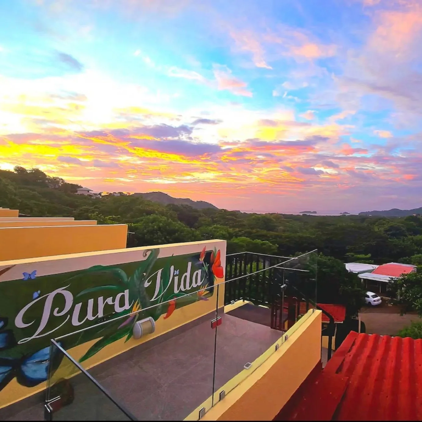 Sunset over a lush green landscape with a colorful sky. In the foreground, there is a rooftop terrace with a sign that reads 'Pura Vida' decorated with butterflies and a hummingbird figure.