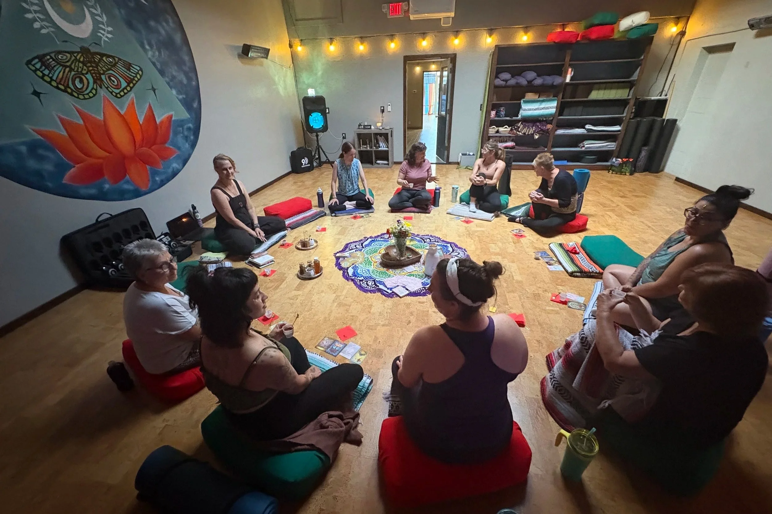 A group of women sitting cross-legged in a circle on the floor participating in a cacao ceremony at a women's circle in a cozy room decorated with string lights and wall art.