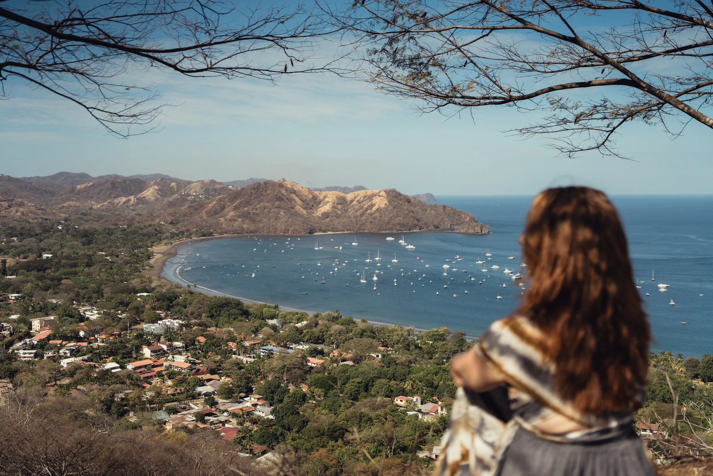 A woman with red hair wearing a patterned scarf looking at a scenic view of the Costa Rican bay with boats, surrounded by hills and trees.