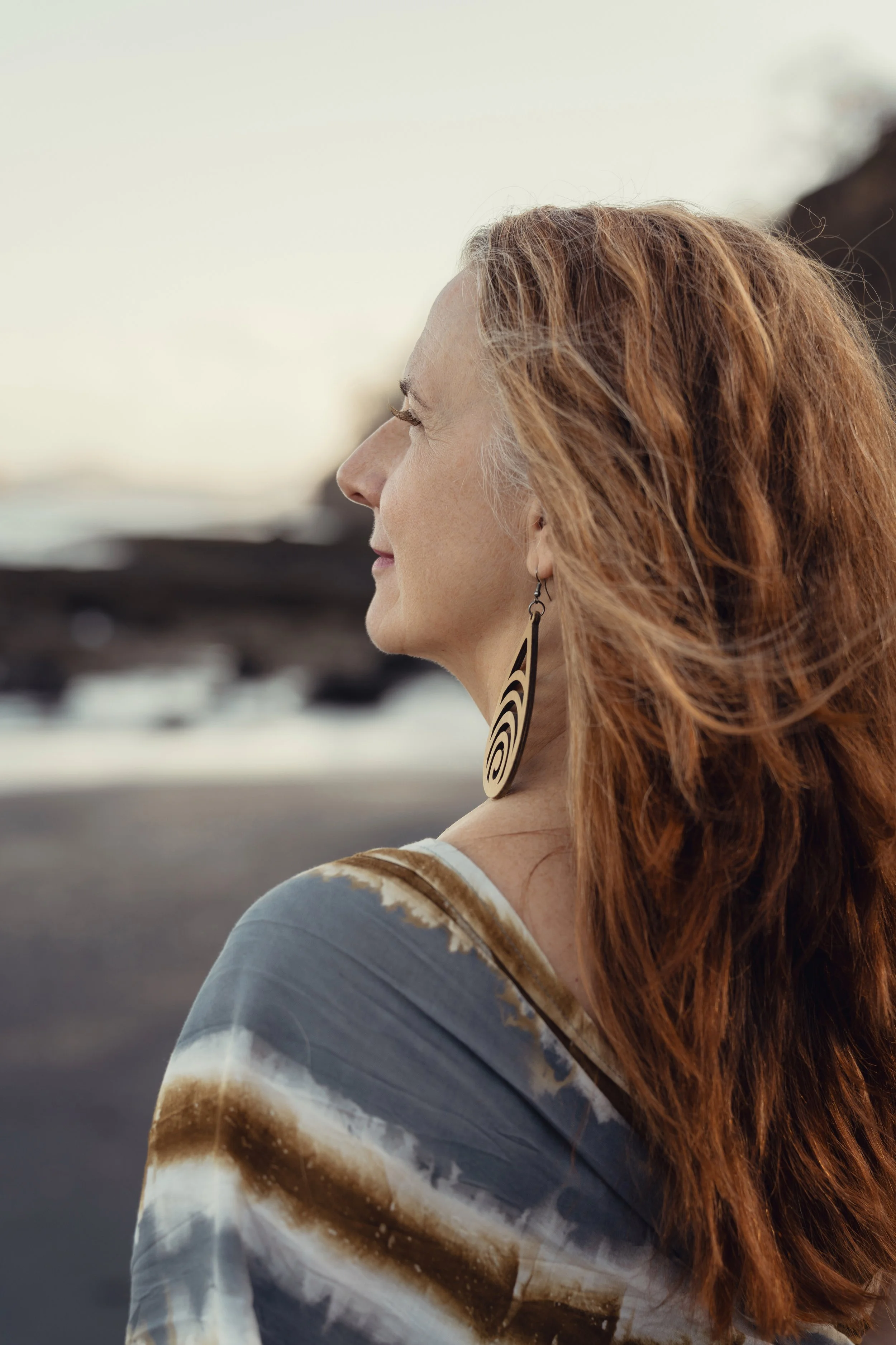 Profile of a woman with long red hair, wearing patterned earrings, standing along the edge of a coast line with a blurred background.