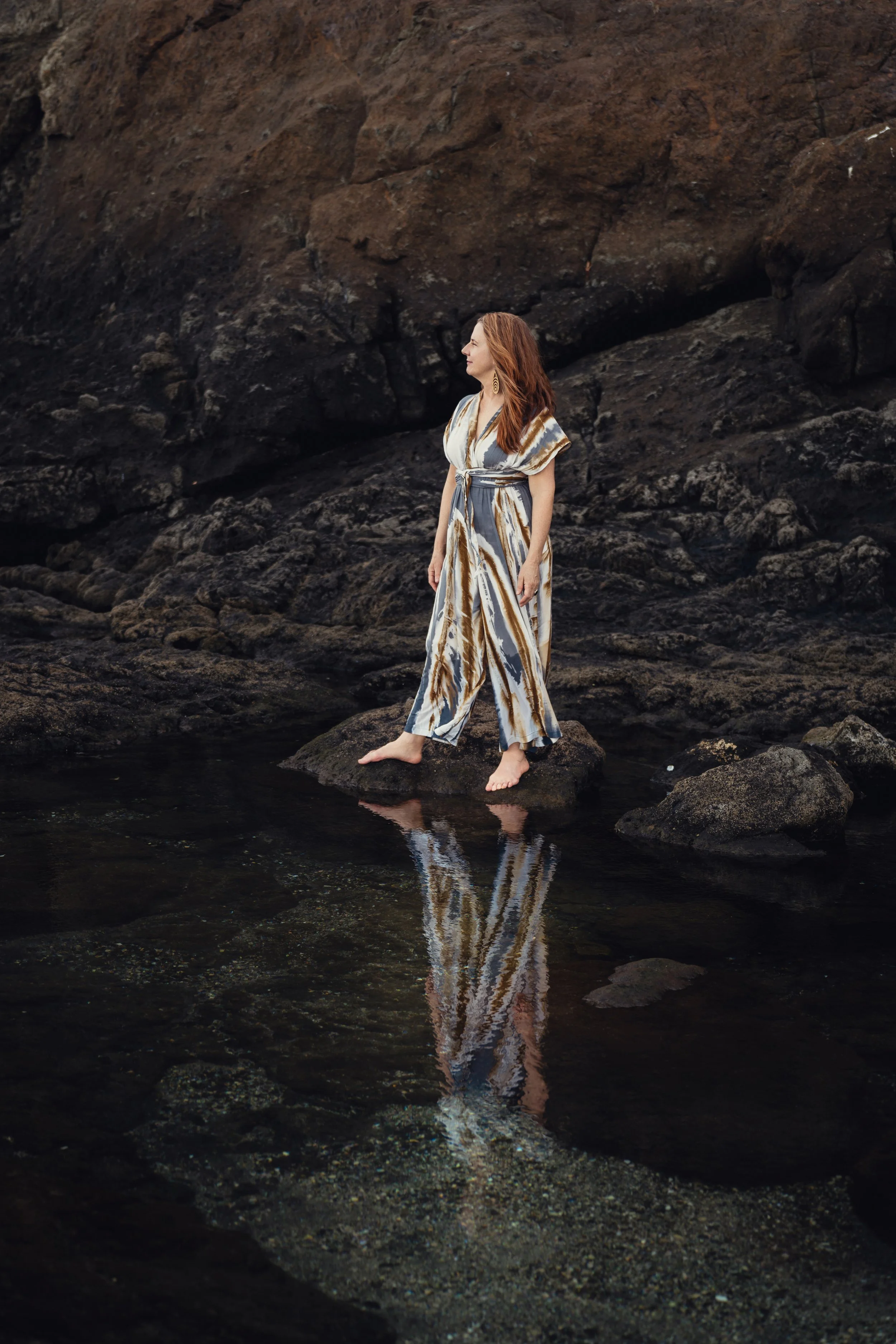 A woman with red hair standing barefoot on a rock in a shallow pool of water at the base of a large, dark rocky cliff.