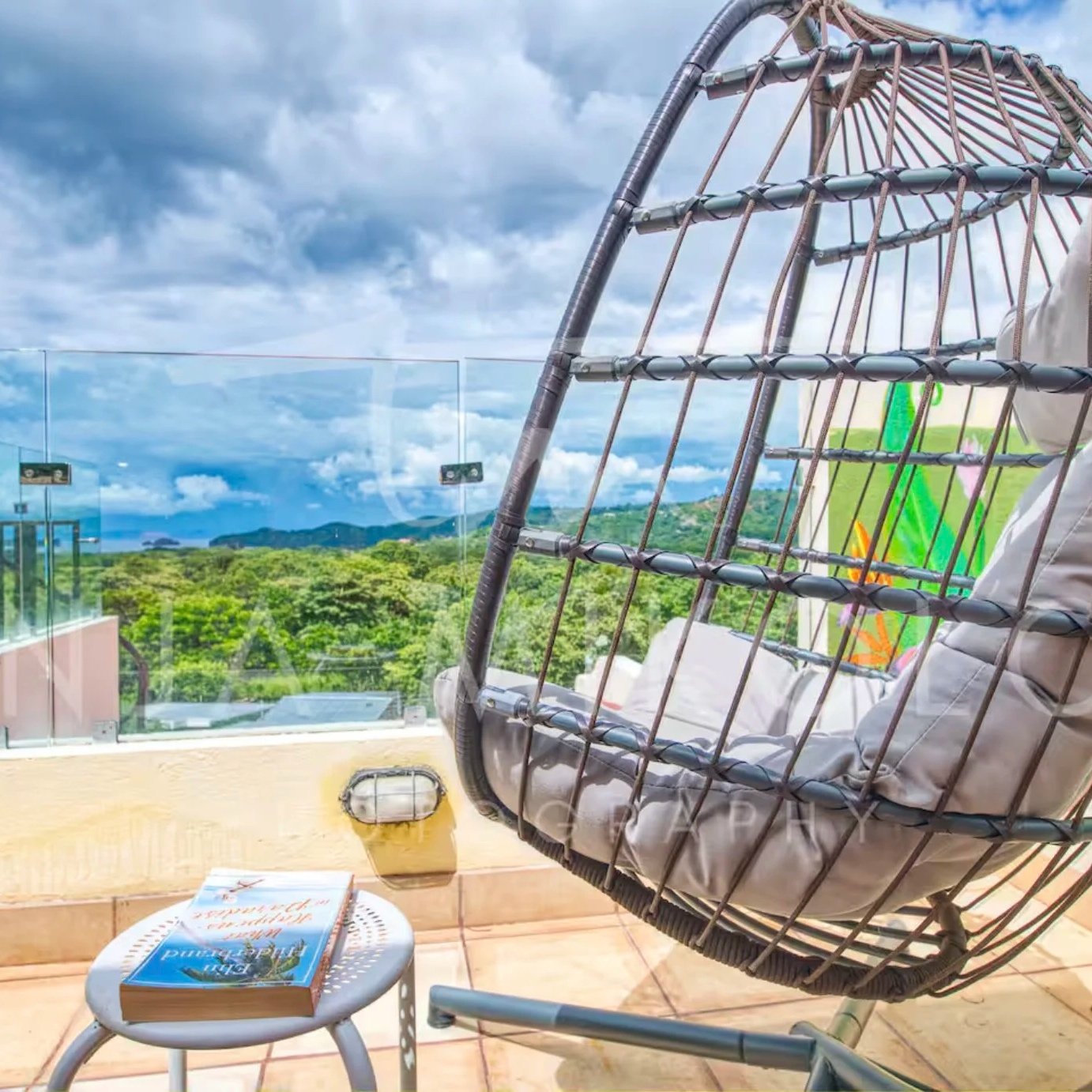 A hanging rattan chair with a cushion on a balcony, overlooking a lush green landscape under a cloudy sky, with glass panels and a small table holding a book.