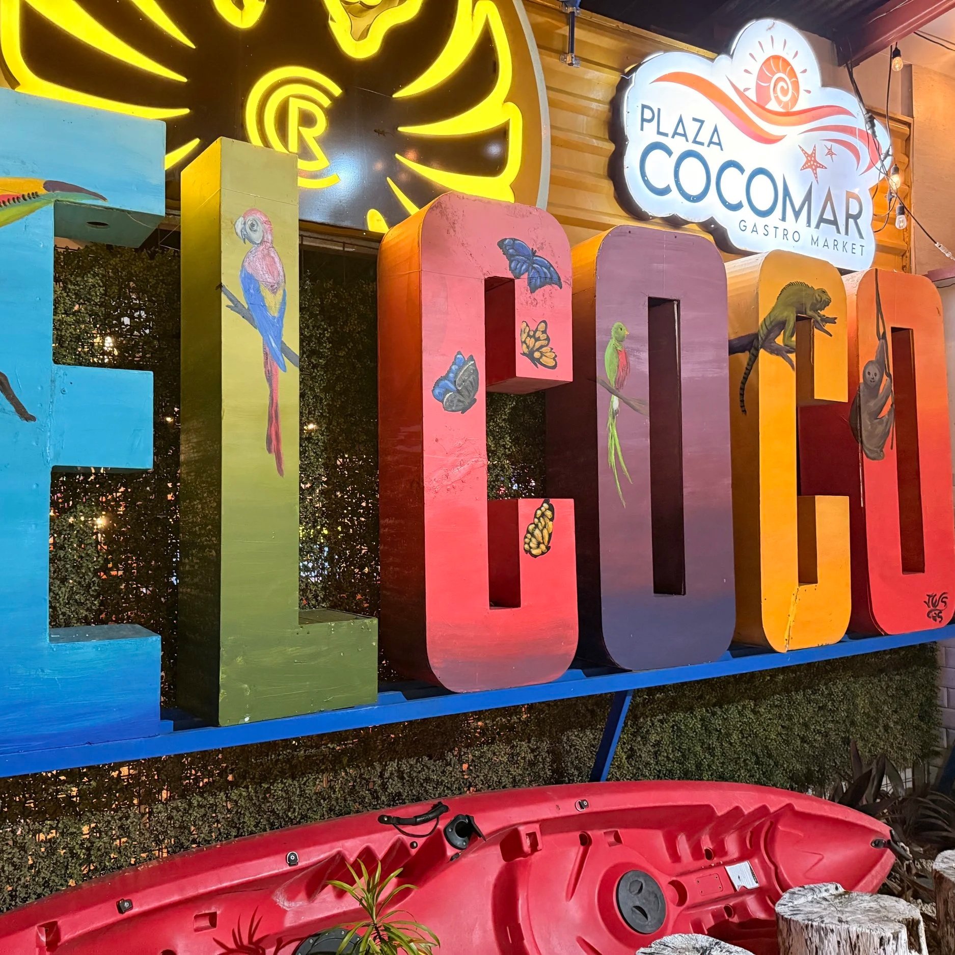 Colorful outdoor sign for Plaza Cocomar Gourmet Market with decorative wood panels painted with animals and butterflies, illuminated signs, and a red canoe at the bottom surrounded by plants and rocks.
