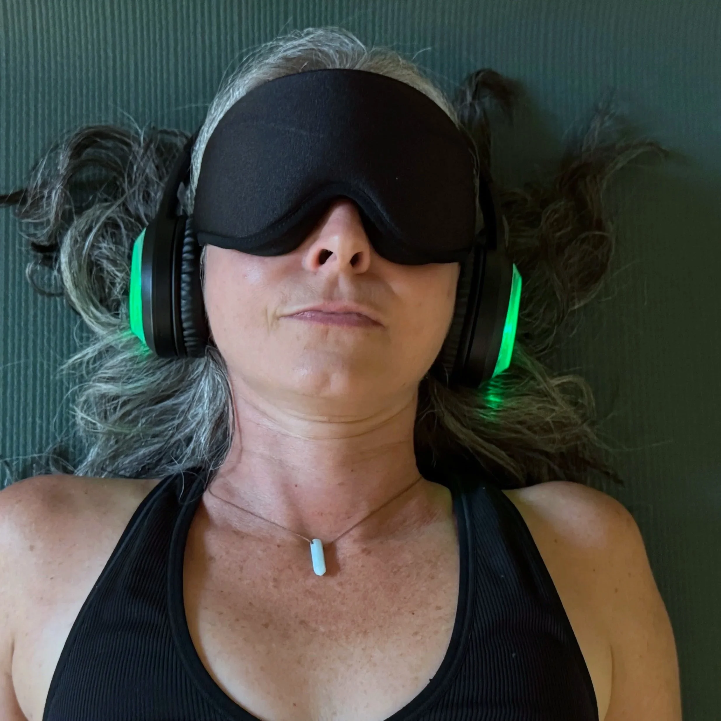 Close-up of a woman with curly gray hair wearing black a eye mask and 9D Breathwork headphones, resting with her eyes closed on a green yoga mat.