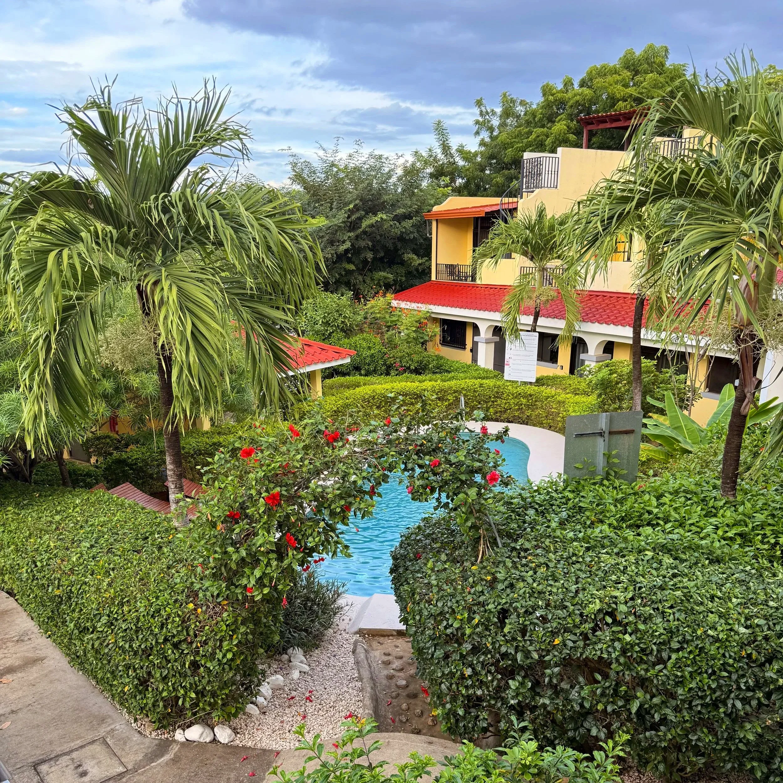 View of a lush, tropical garden with palm trees, hibiscus flowers, and dense green foliage surrounding a yellow house with red tile roof and small balcony, under a partly cloudy sky.