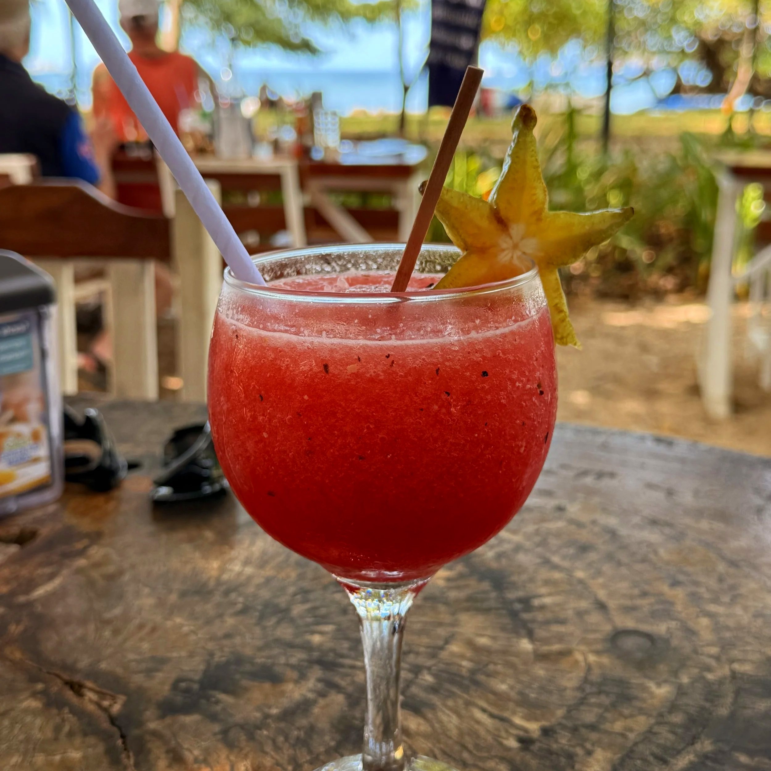 Close-up of a red beverage in a glass, garnished with a starfruit slice, two straws, and condensation on the glass. Blurred outdoor setting in the background.