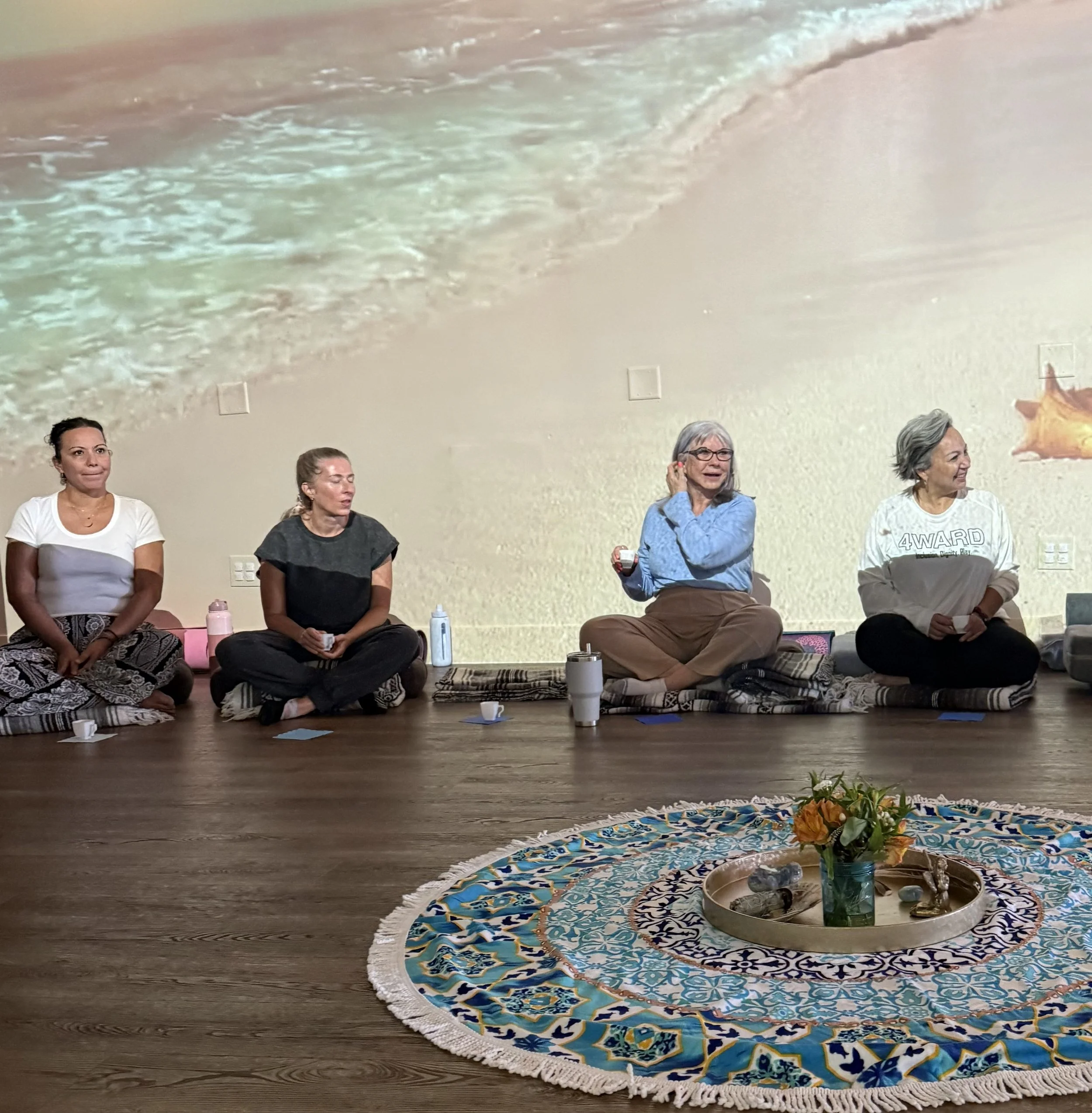 A group of people sitting on cushions in a circle on a wooden floor, participating in a discussion, with a decorative rug and flowers in the foreground.