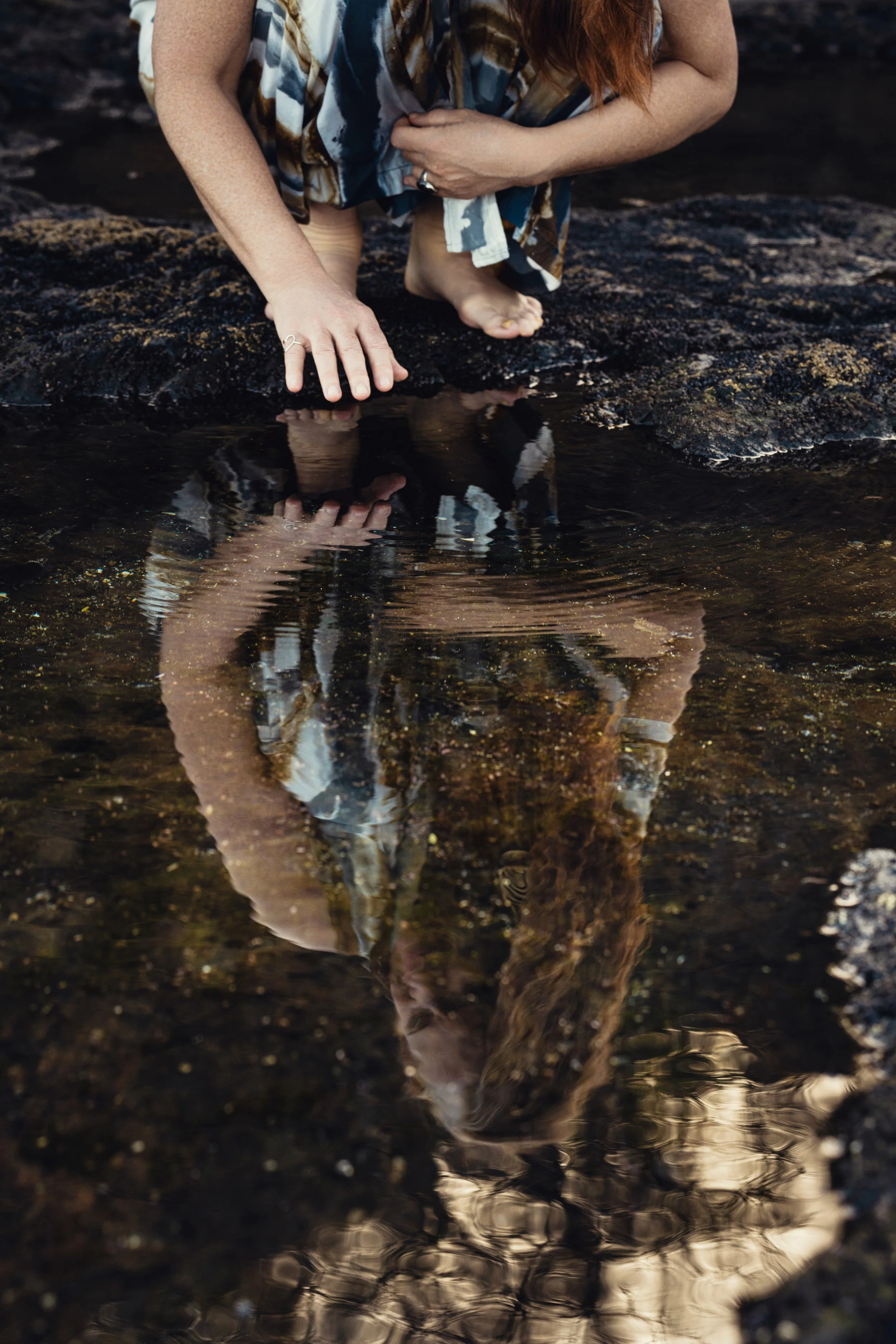 A woman with red hair crouching near shallow water in tidal pools on a rocky surface, touching the water with their left hand. The reflection of the woman and surrounding rocks can be seen in the water.
