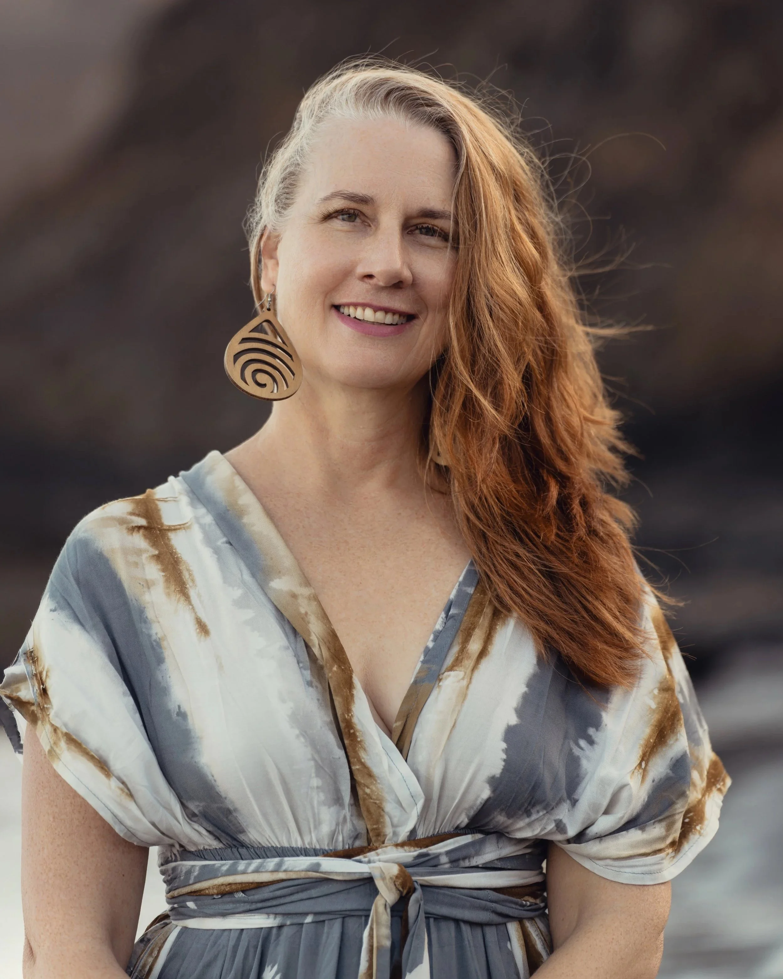 A woman with long red hair, smiling, wearing a large wooden earring and a tie-dye style dress with beige, gray, and white colors, standing outdoors with a blurred background.
